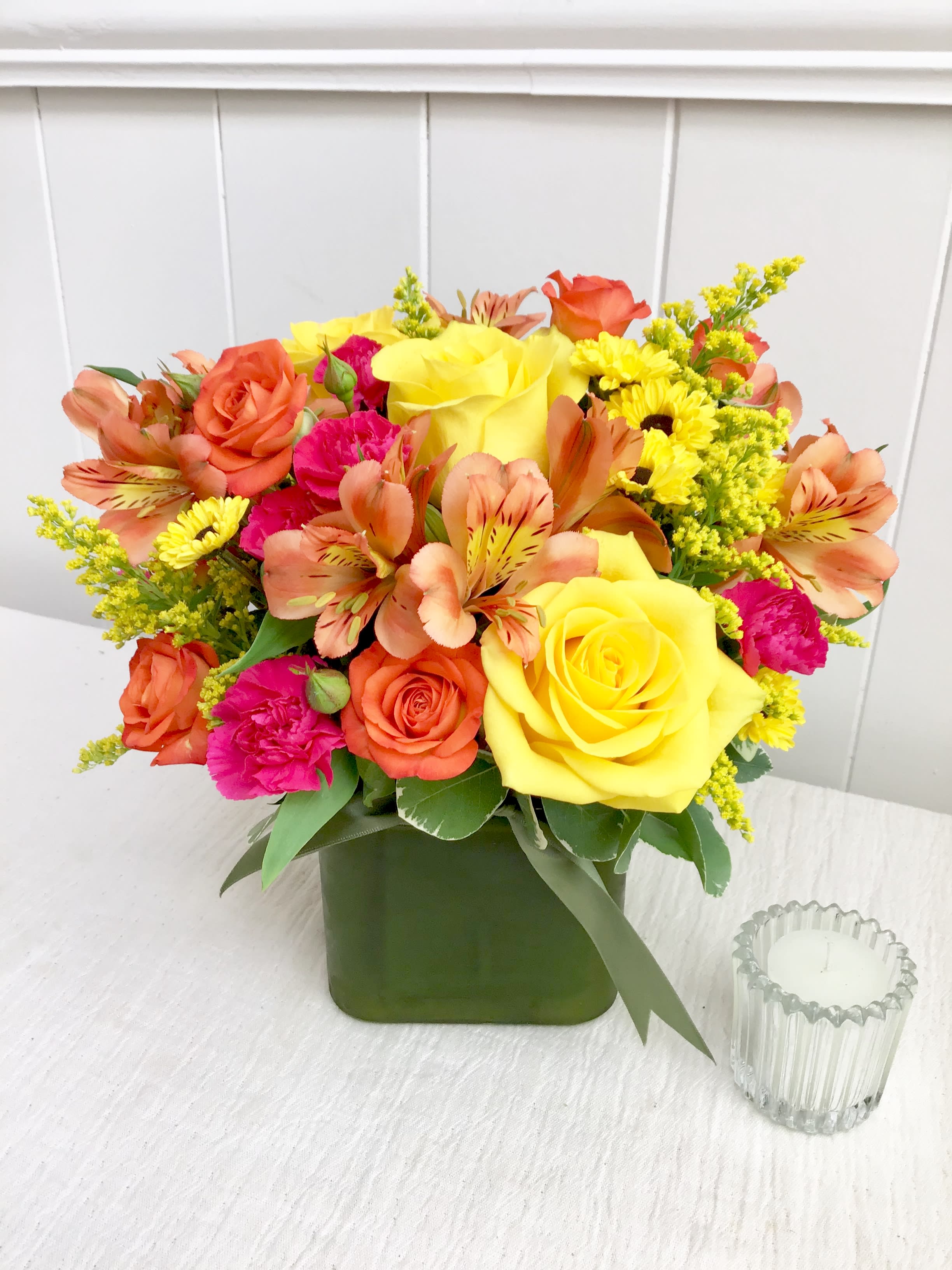 Bright arrangement of yellow roses, orange alstroemeria, and pink blooms in a green cube vase.