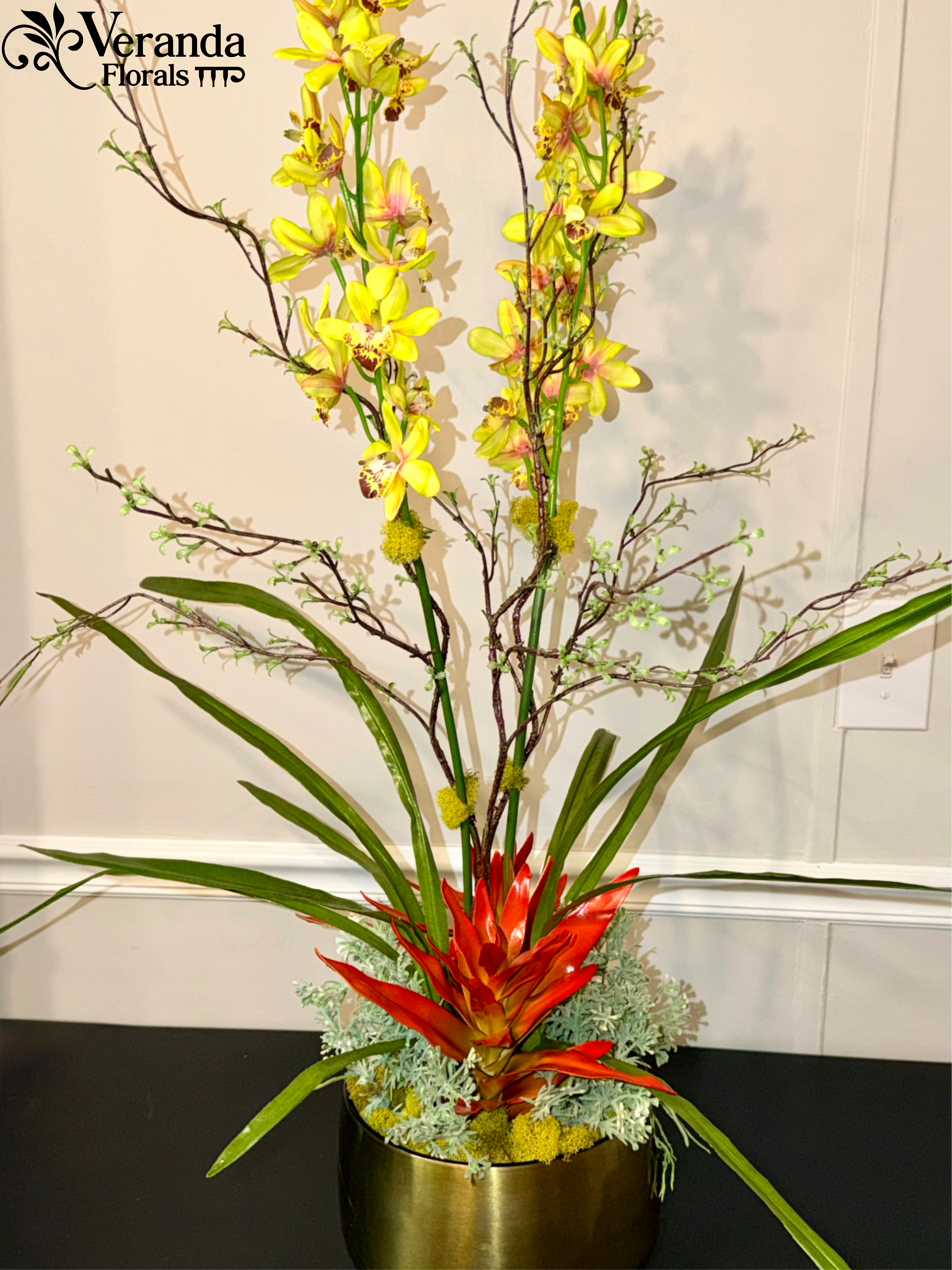 Tall tropical arrangement with yellow orchids and red bromeliad in a gold pot