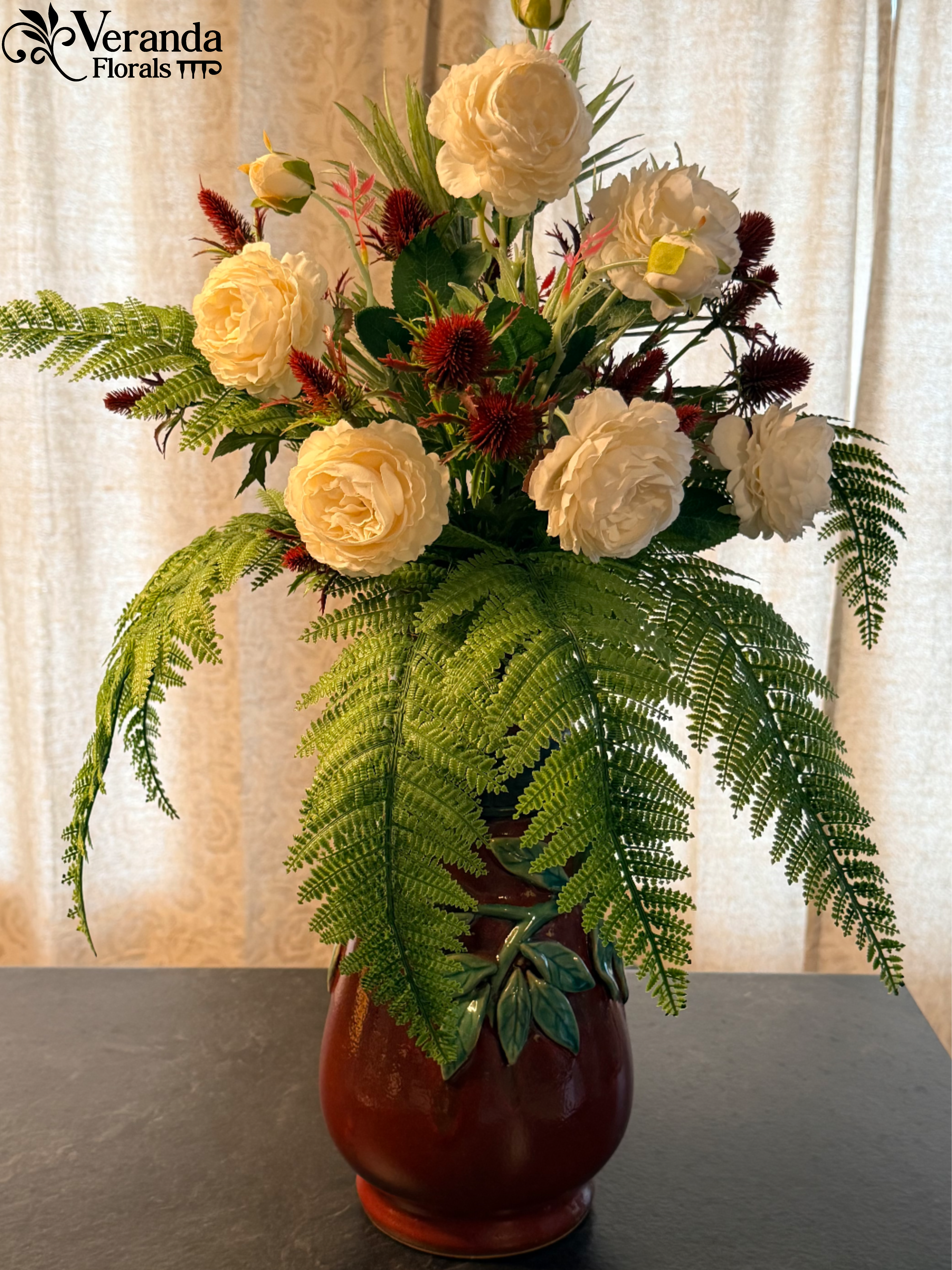 Tall arrangement of cream blooms with red accents and cascading fern fronds in a red ceramic vase