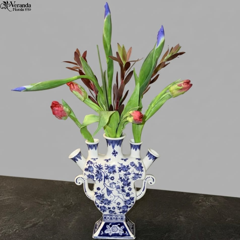 Blue and White Tulipiere - Elevate your floral design with this elegant Blue and White Tulipiere. Inspired by classic Delftware, this vessel features a central spout and four individual side ports, allowing for effortless, structural arrangements. Whether featuring delicate spring bulbs or architectural branches, its unique silhouette makes it a statement piece even when empty. Price includes the vase and designers choice of flowers. 