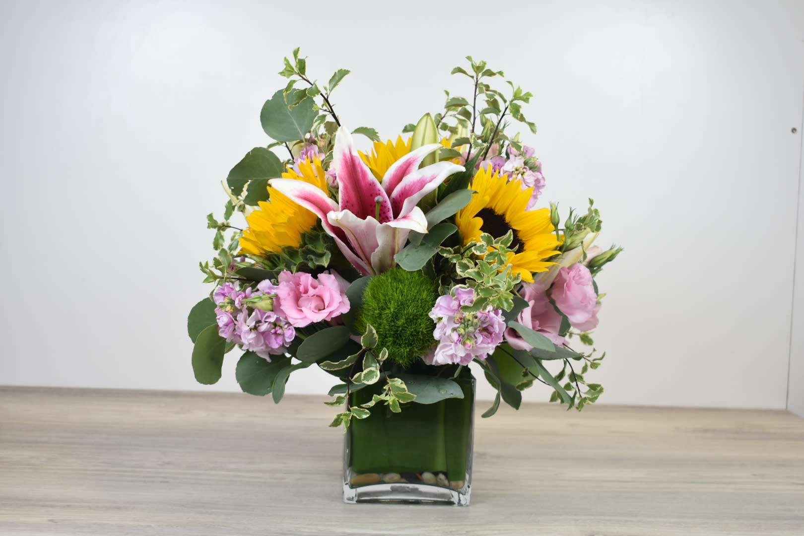 Bright mixed arrangement with sunflowers, pink lilies, and pastel blooms in a square glass vase