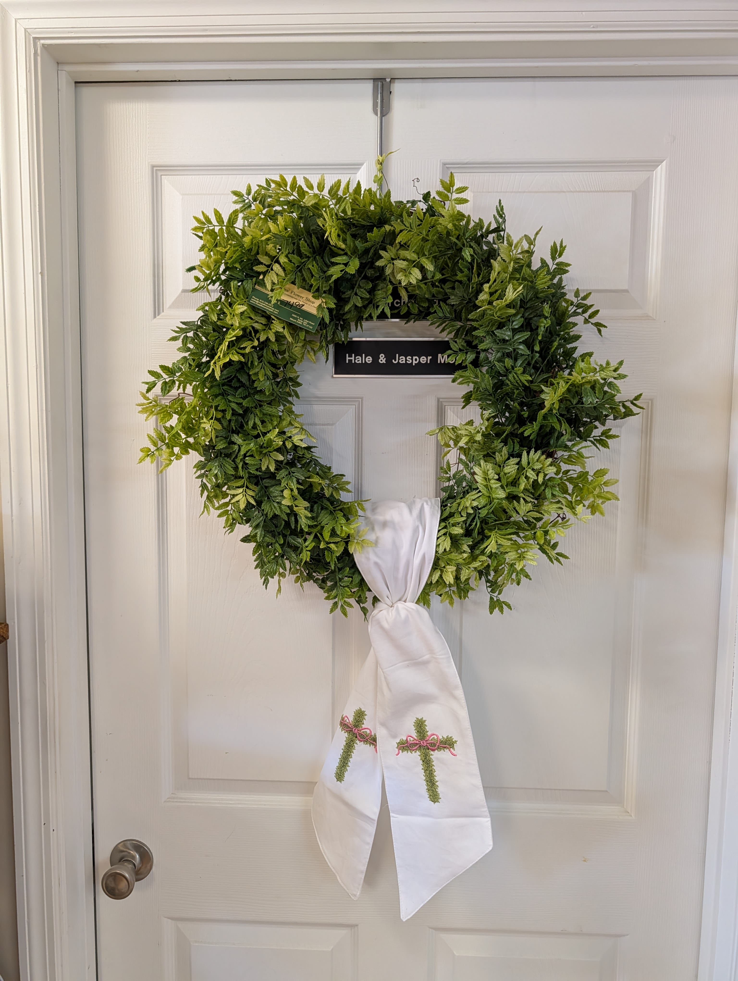 Graceful Greenery Cross Wreath - A simple, reverent accent for any season, this permanent botanical wreath features lush all‑green foliage finished with a white sash tie embroidered with delicate crosses. Clean and understated in style, it’s a beautiful way to add a touch of faith and serenity to your front door, mantle, or memorial display all year long. 