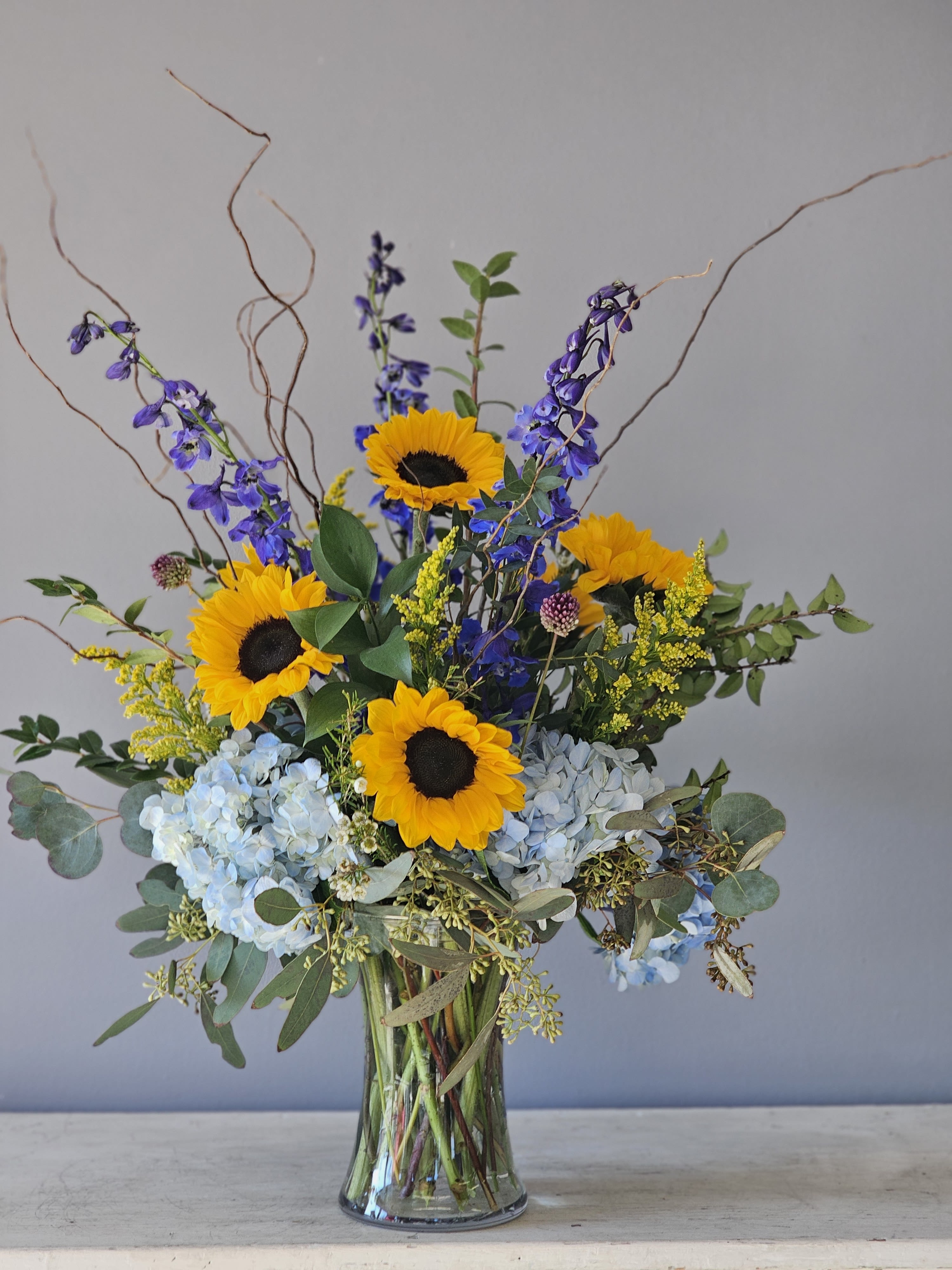 Tall arrangement of yellow sunflowers, blue hydrangeas, and delphinium in a clear glass vase