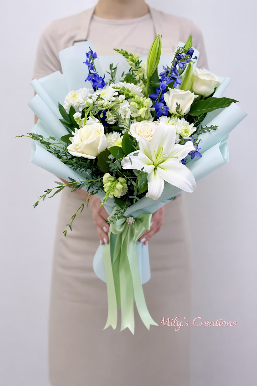 Hand-tied bouquet of white lilies, roses, and blue flowers wrapped in pale blue paper with green ribbon.