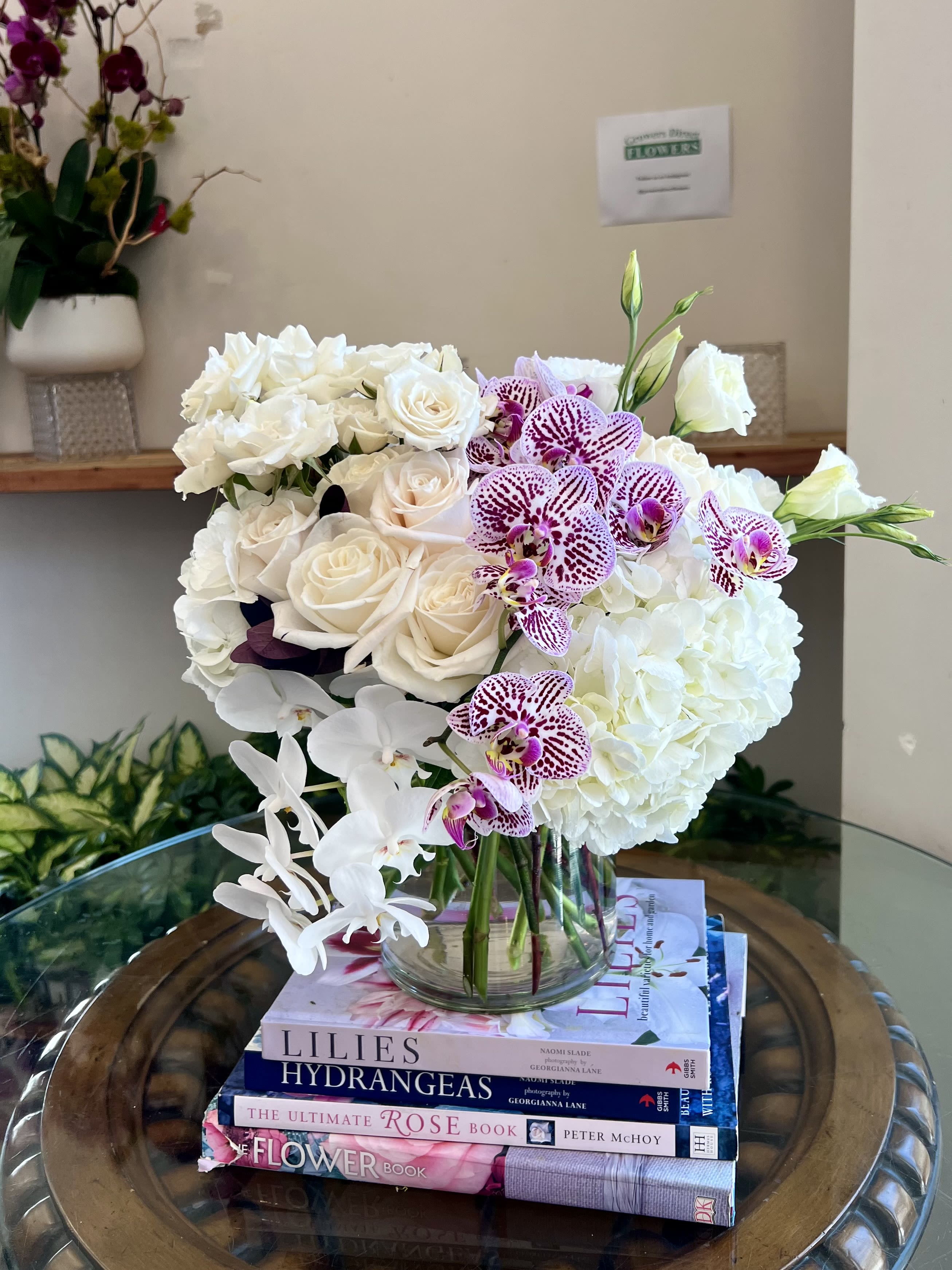 White roses, hydrangeas, and cascading orchids with purple speckled blooms in a clear glass vase on stacked books.