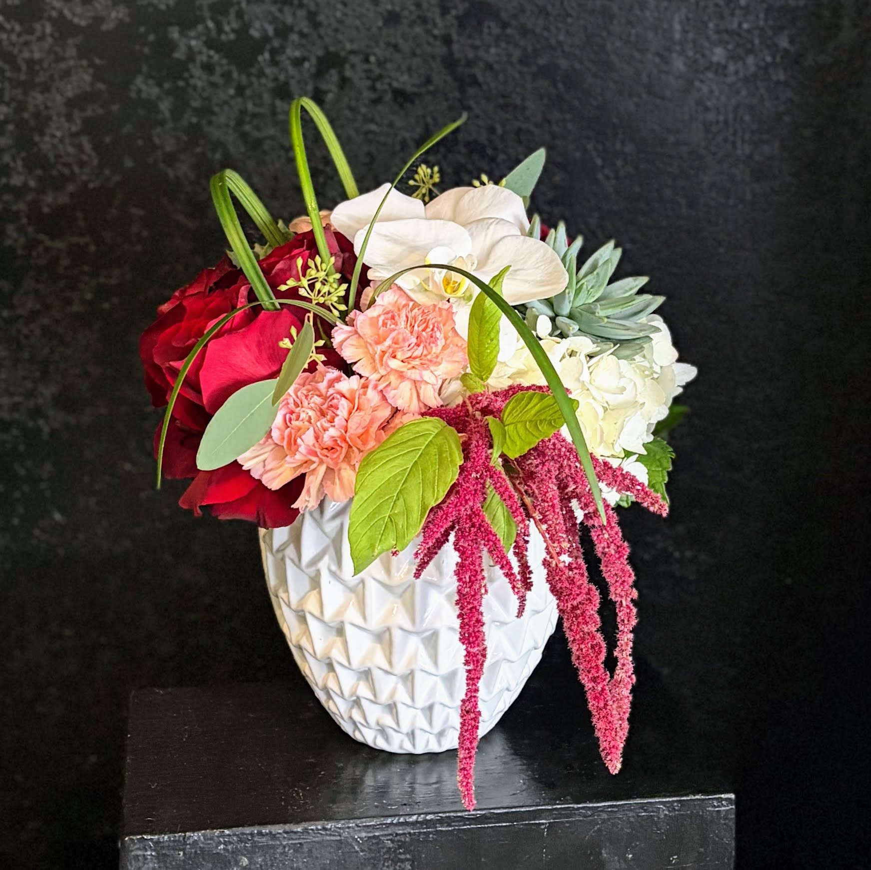 Asymmetrical mixed arrangement with red roses, peach carnations, white orchids, and succulents in a white ceramic vase