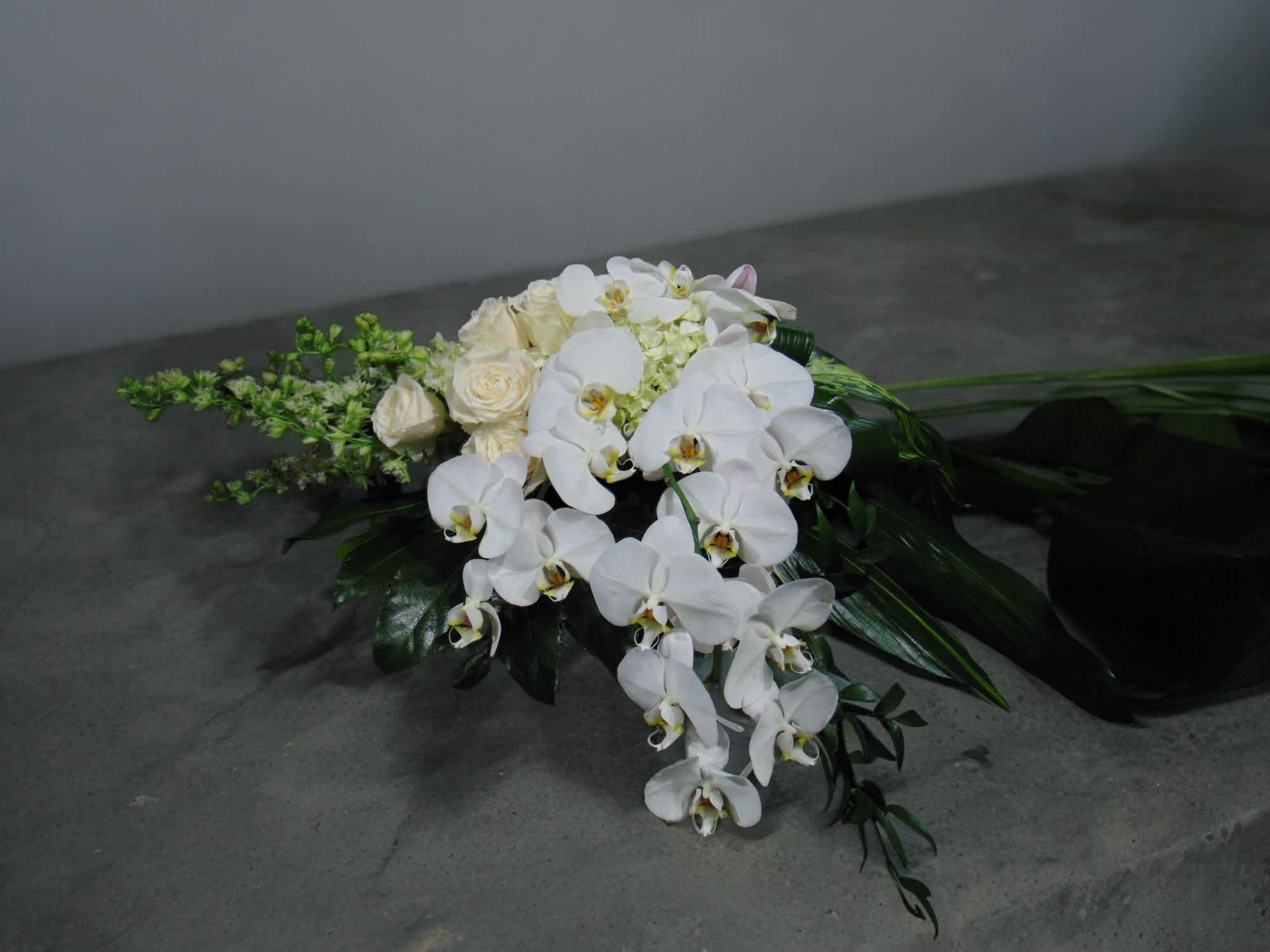 Cascading bouquet of white orchids and ivory roses with dark tropical foliage on a gray surface