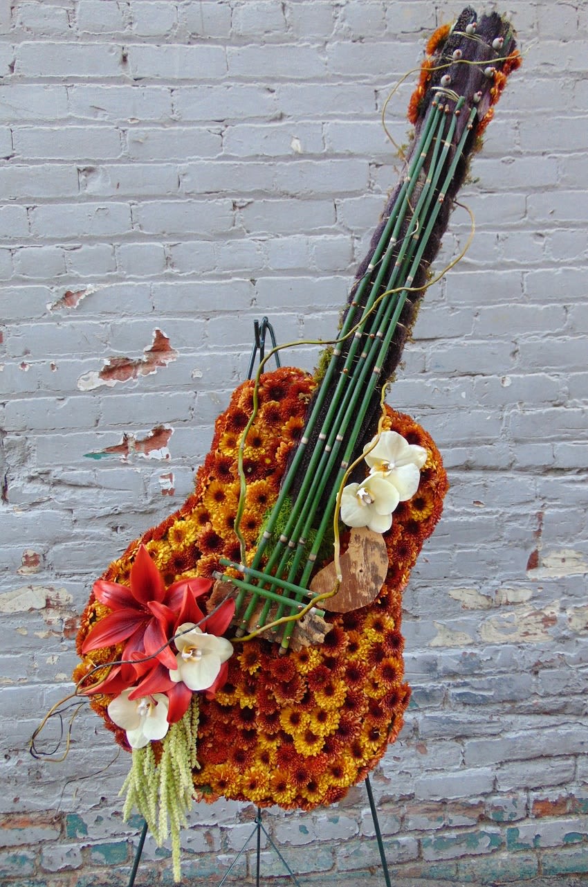 Tall floral arrangement shaped like a guitar with orange mums, red lilies, and white orchids on an easel.