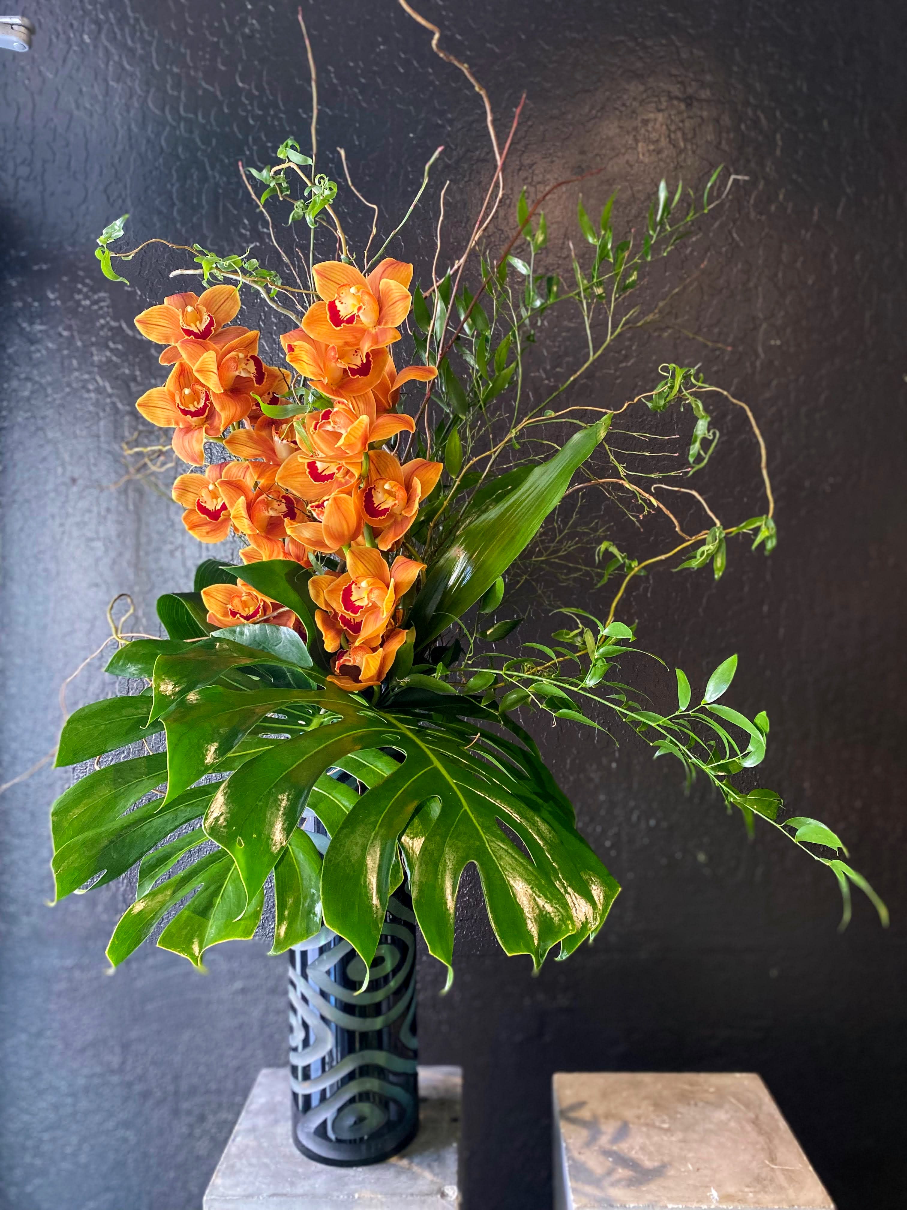Tall arrangement of orange orchids with large monstera leaves in a patterned black cylinder vase