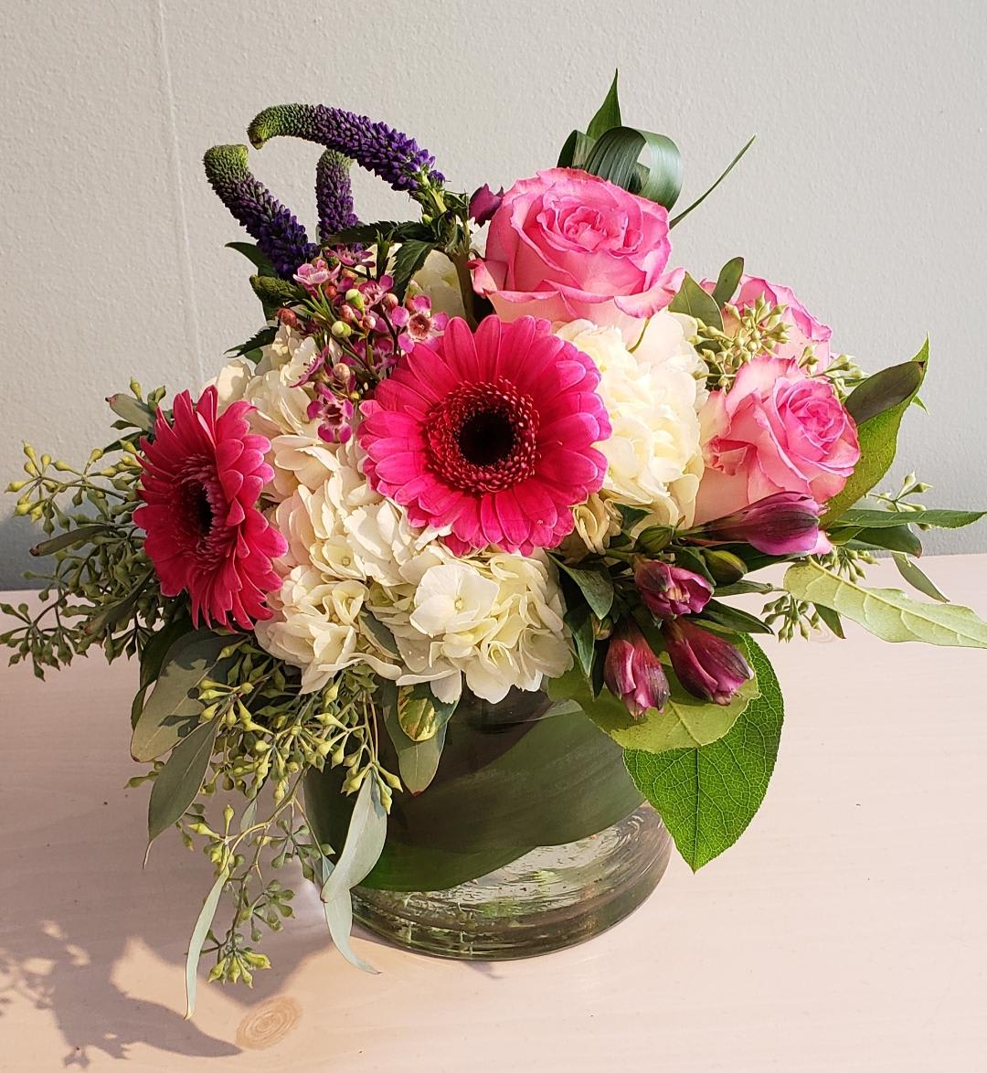 Low round arrangement of pink gerbera daisies, pink roses, and white hydrangeas in a clear glass vase