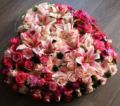 Heart-shaped arrangement of pink lilies, roses, and gerbera daisies in various pink and red tones