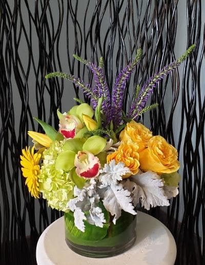Compact yellow and green floral arrangement with orchids, roses, hydrangea and purple accents in a glass vase