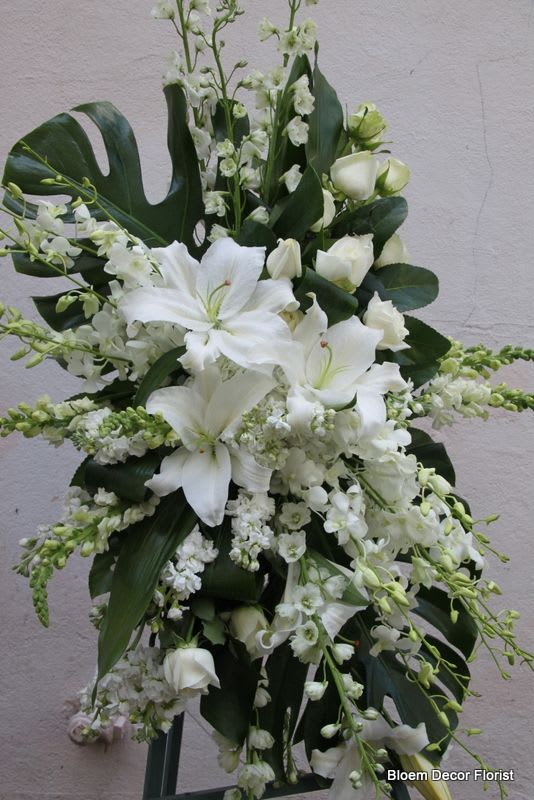 Tall standing spray of white lilies, roses, and other white flowers with large dark tropical leaves.