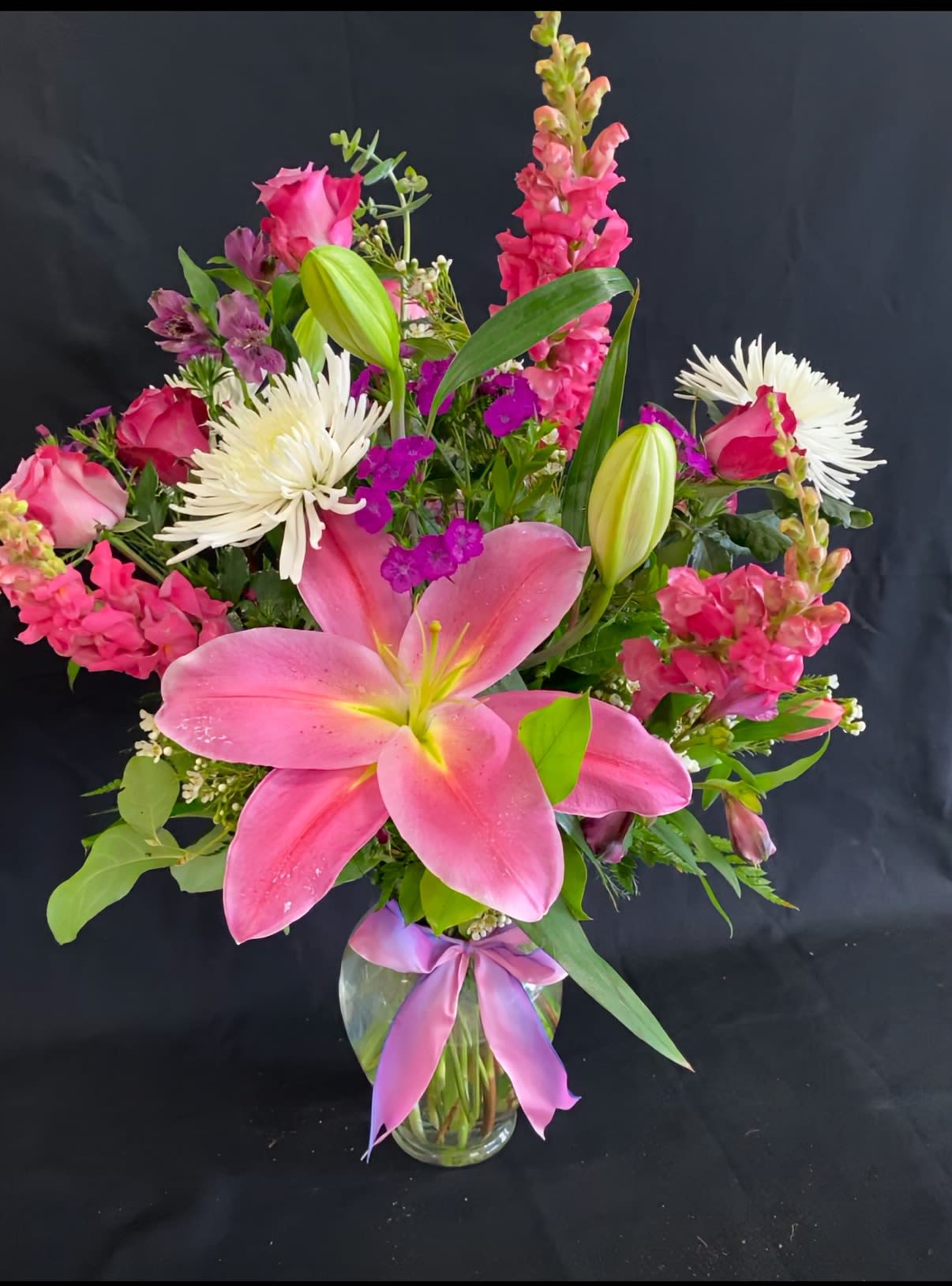Tall mixed arrangement of pink lilies, roses, snapdragons and white mums in a clear vase with a ribbon bow