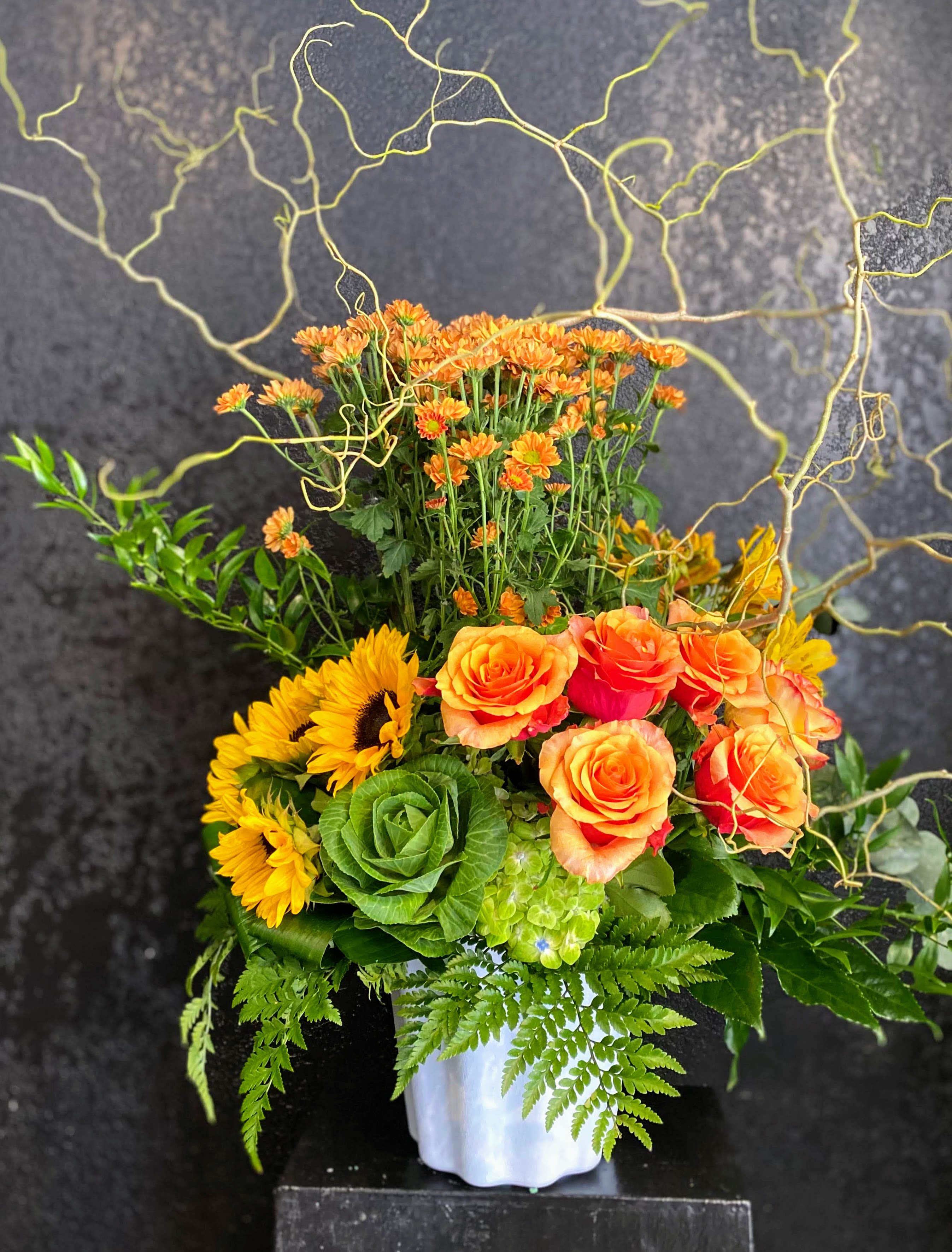 Tall orange and yellow arrangement with roses, sunflowers and mums in a white vase with curly branches