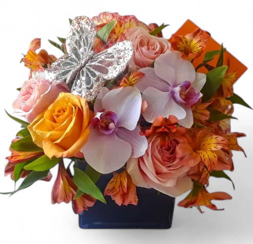 Compact arrangement of roses, orchids, and alstroemeria in a blue cube vase with a silver butterfly decoration
