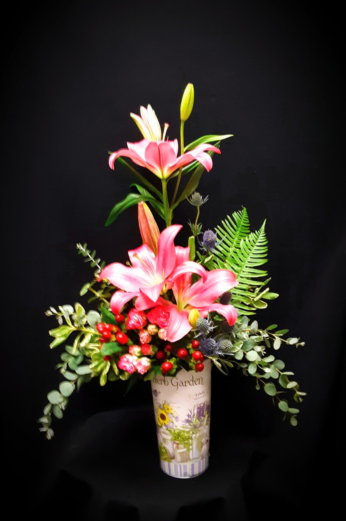 Tall arrangement of pink lilies, small roses, and red berries in a decorative herb garden vase