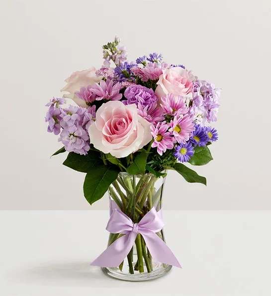 Pastel pink and purple roses and daisies arranged in a clear vase with a lavender bow