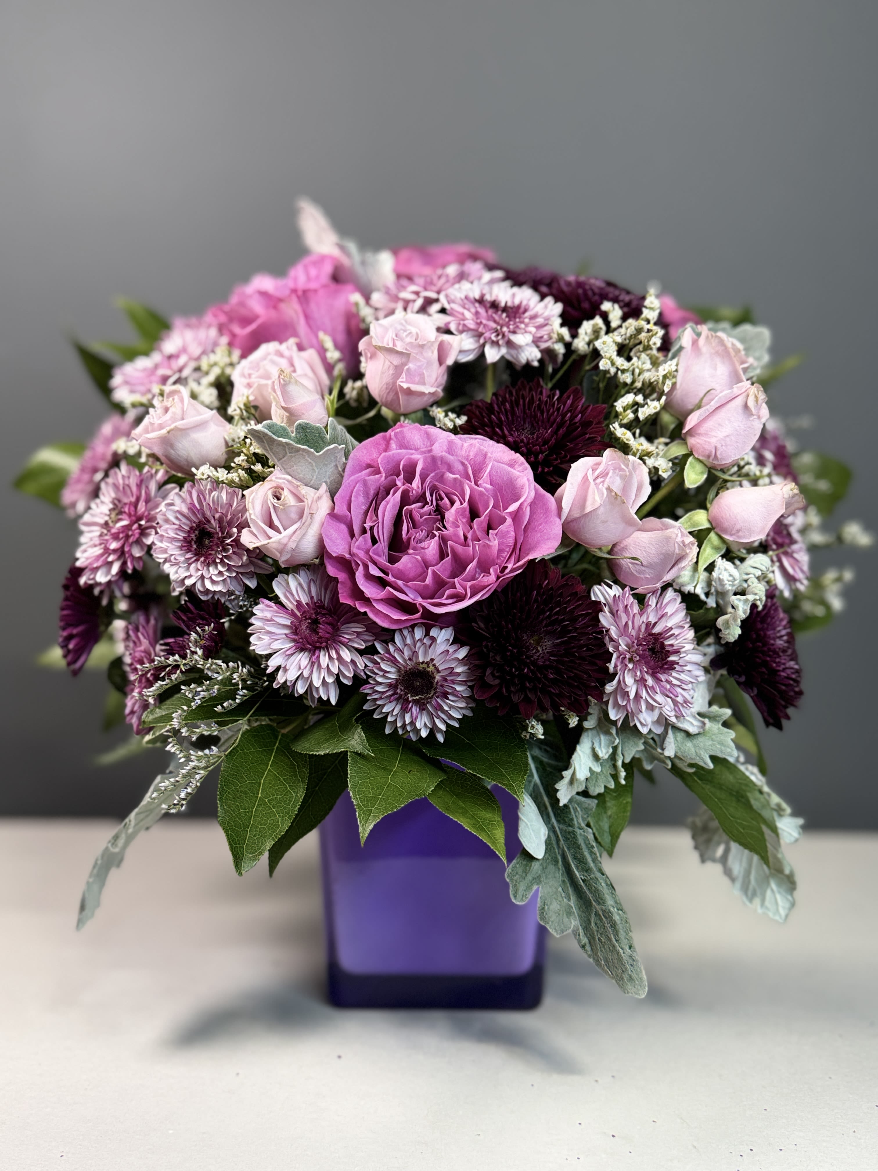 Rounded arrangement of pink roses and purple mums in a purple cube vase