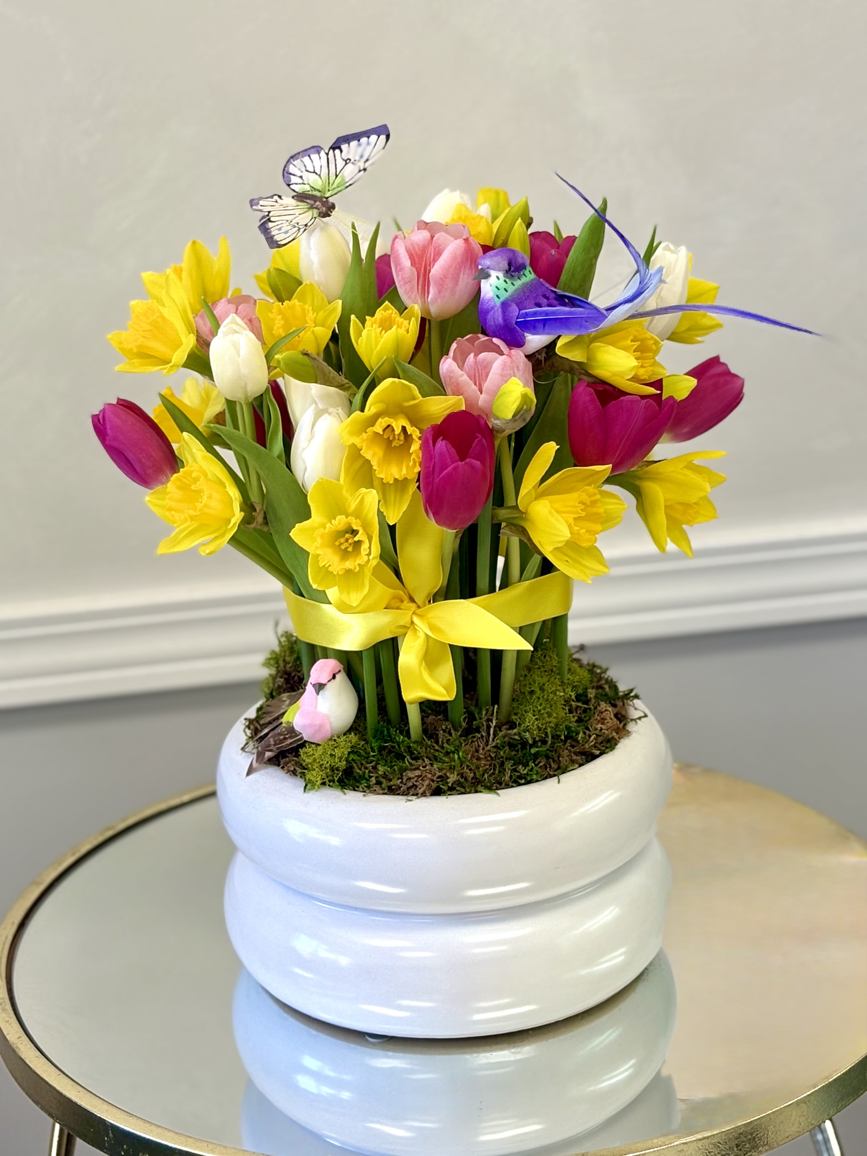 Bright arrangement of yellow daffodils and multicolor tulips in a white pot with decorative birds and butterfly