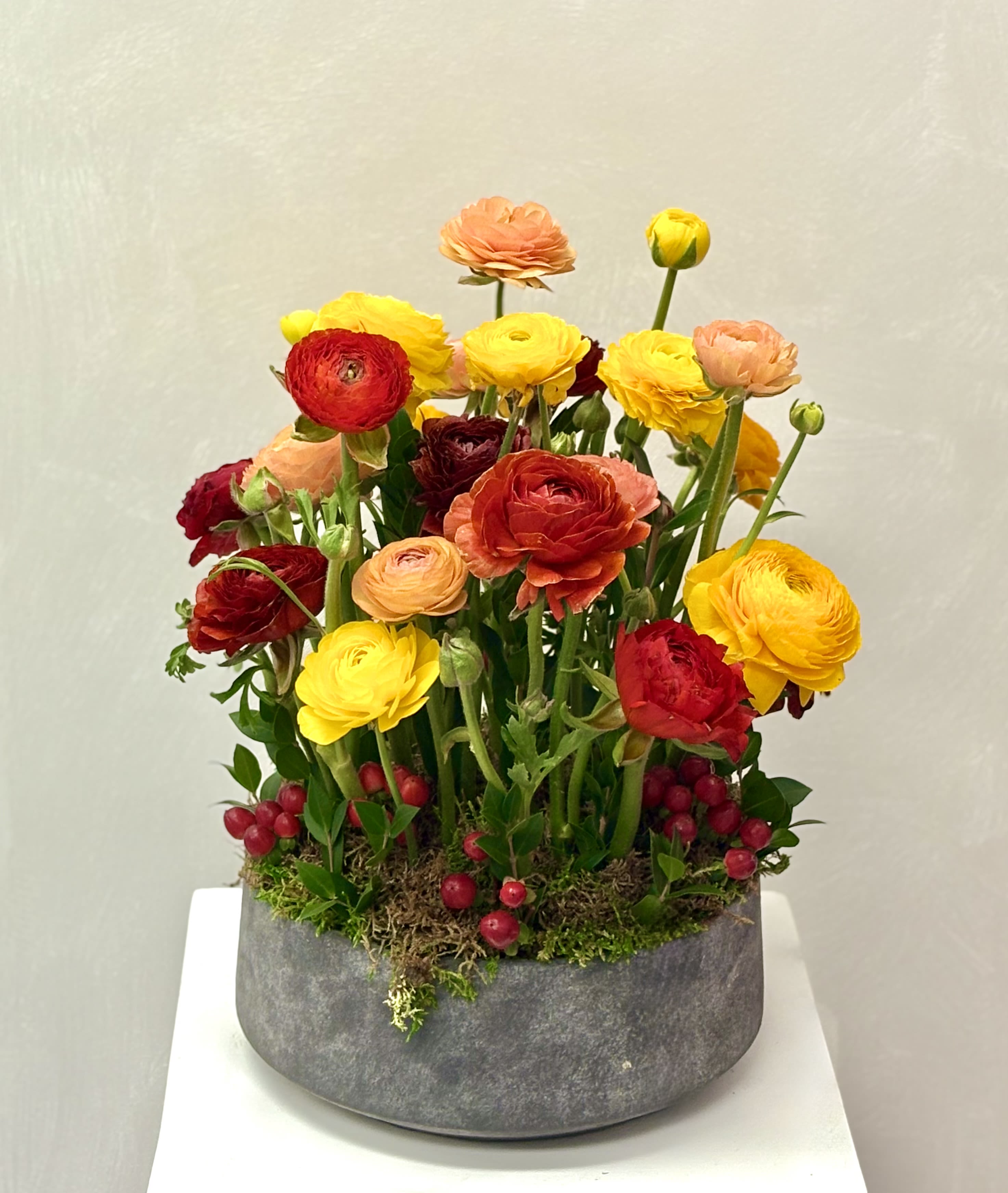 Low gray bowl filled with tall yellow, orange, and red blooms accented with red berries.