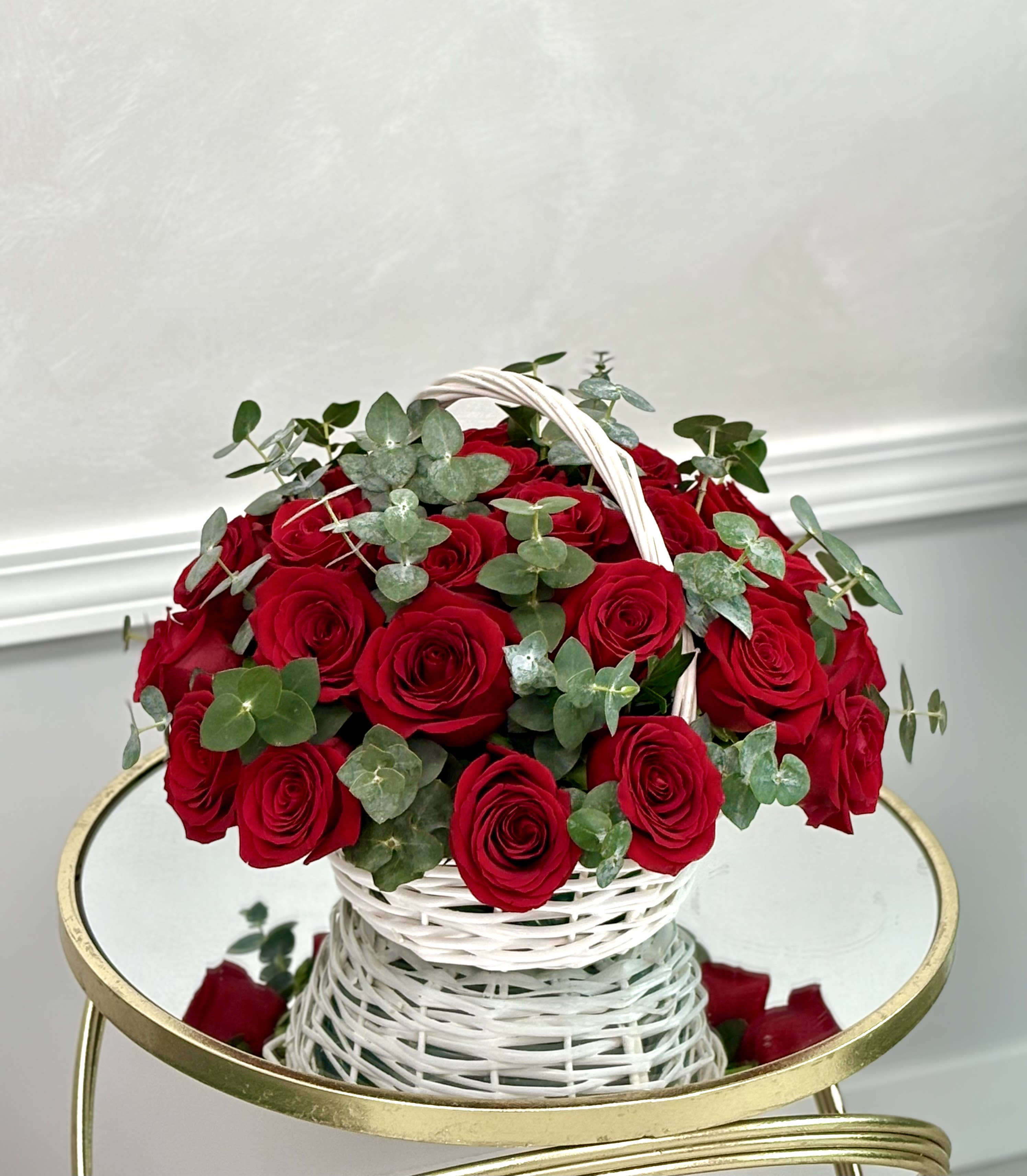 Round basket of red roses with eucalyptus in a white woven handle basket on a mirrored table