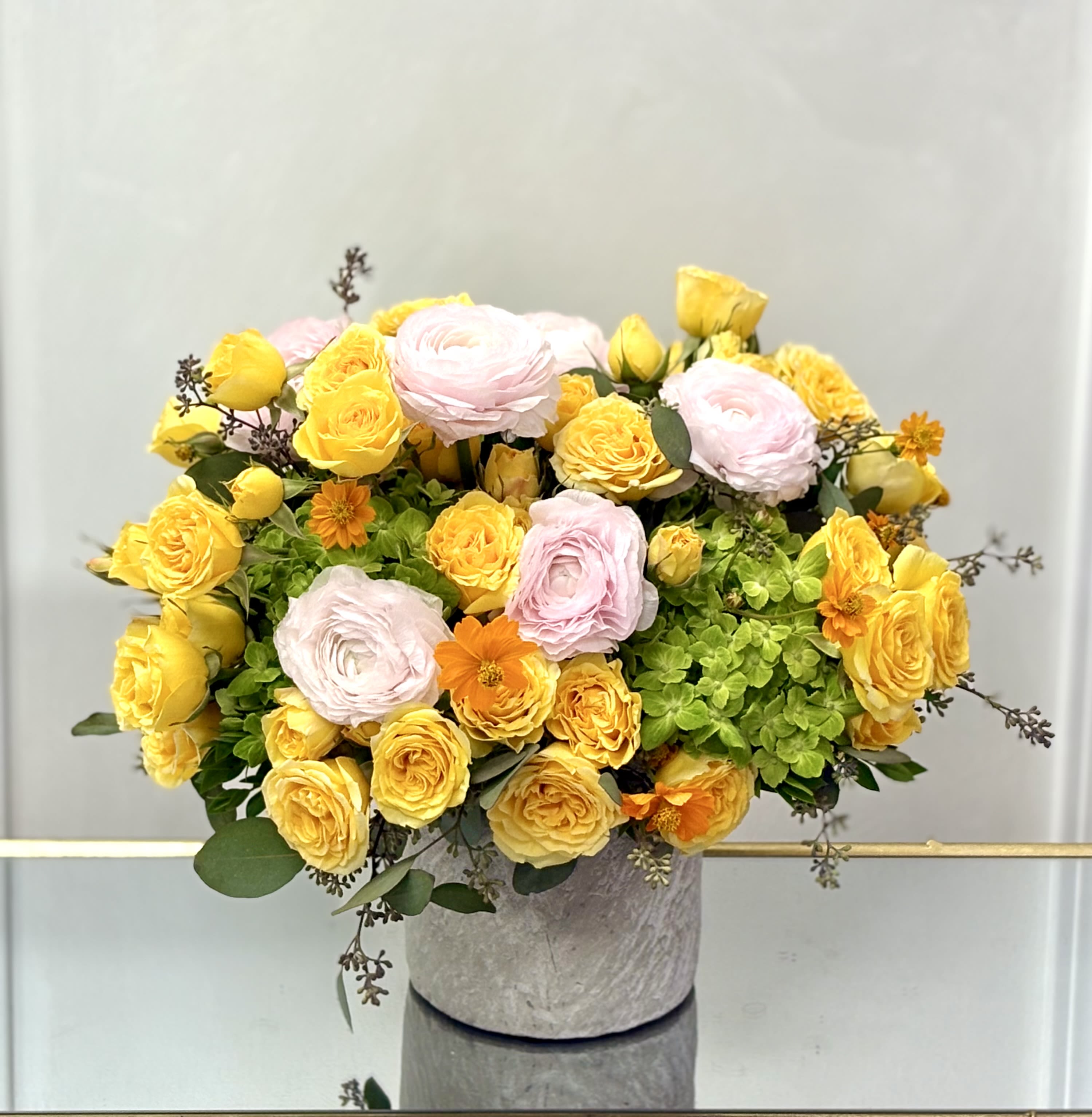 Yellow roses and soft pink blooms arranged with green hydrangeas in a round gray vase.