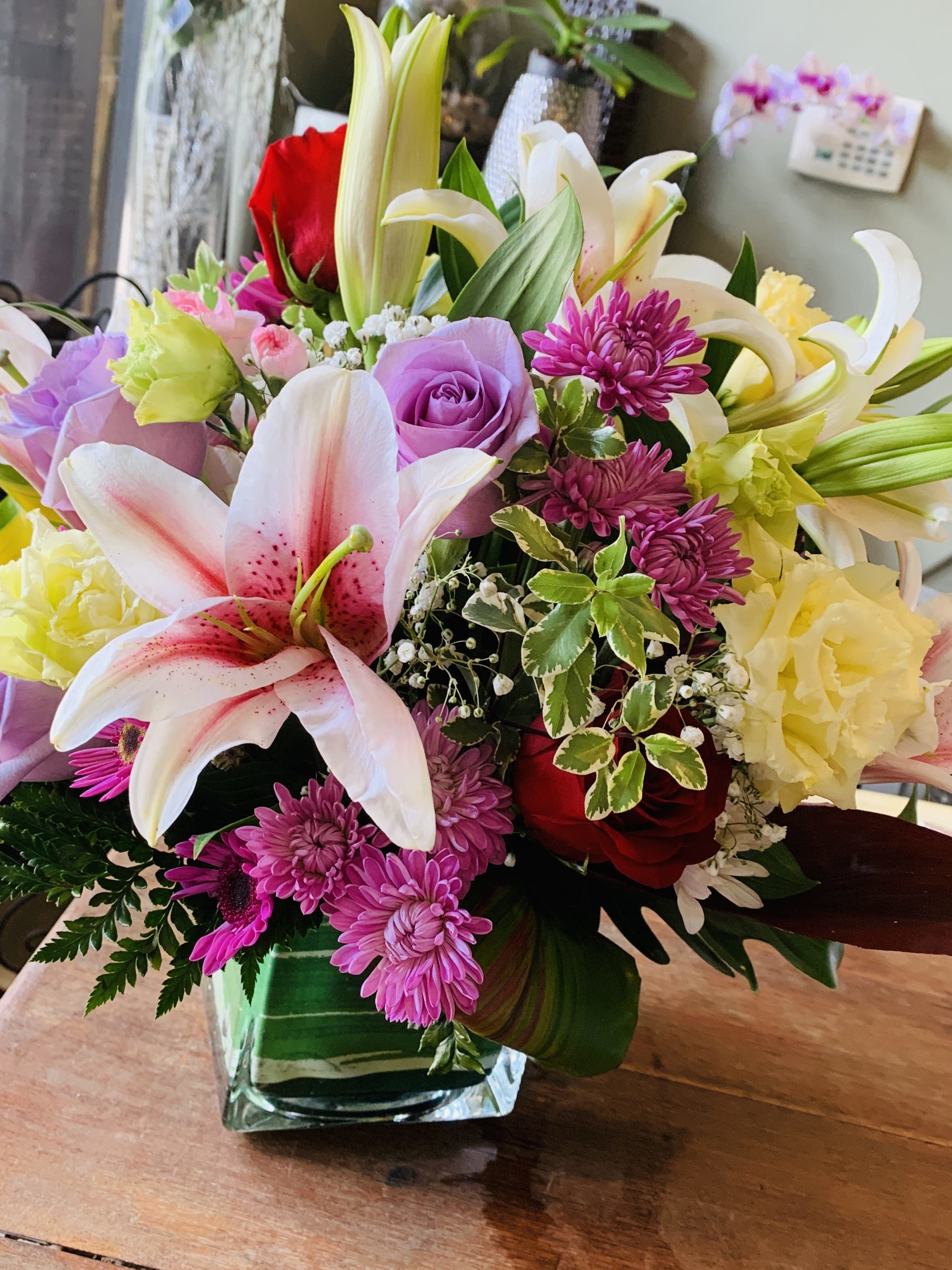 Mixed arrangement of pink lilies, purple roses, red roses, and yellow blooms in a square glass vase