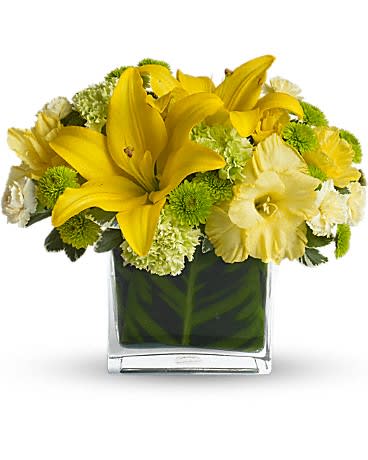 Low arrangement of yellow lilies and mixed yellow and green flowers in a clear glass cube vase
