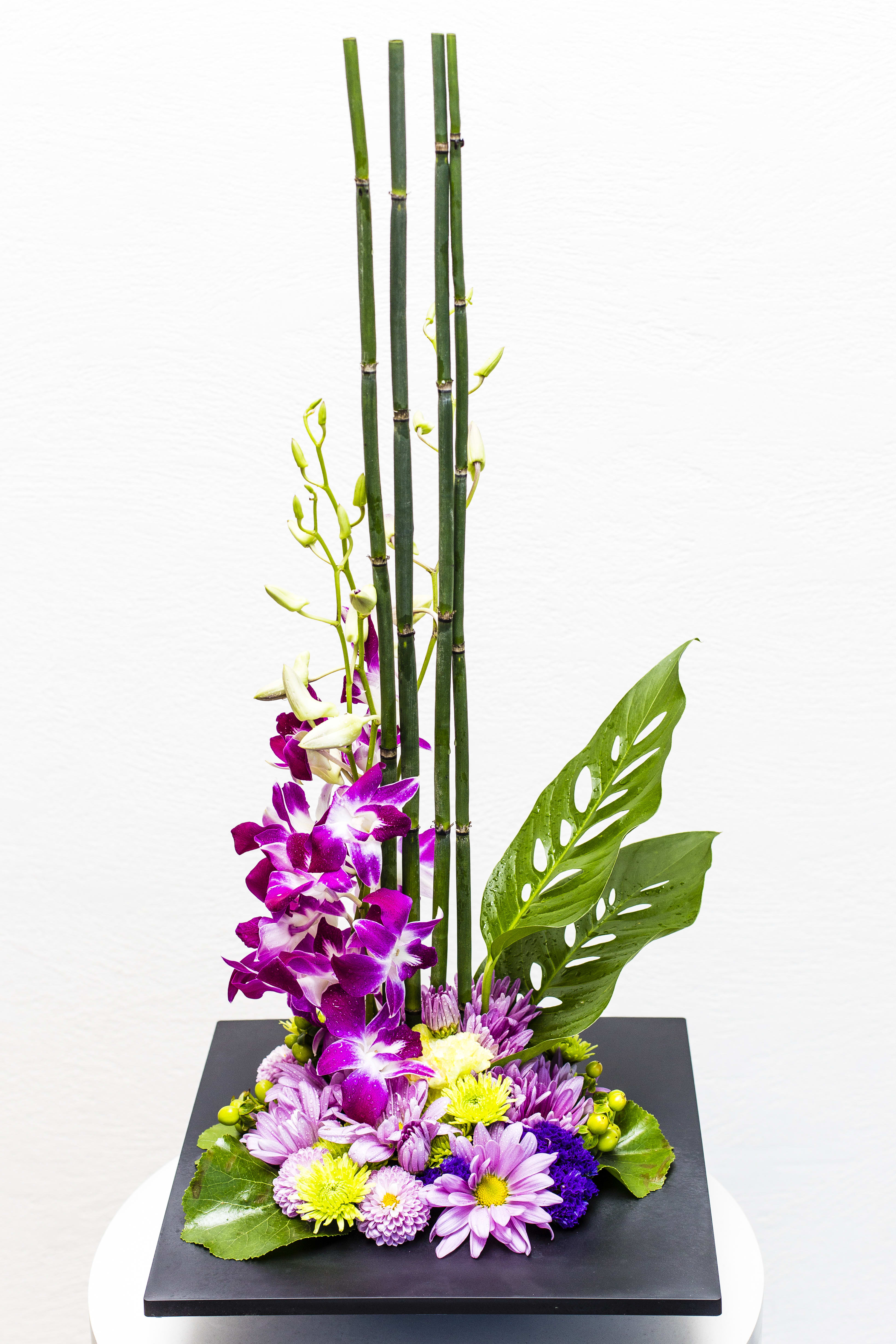 Tall modern arrangement with purple dendrobium orchids and mixed mums around bamboo in a black square base.