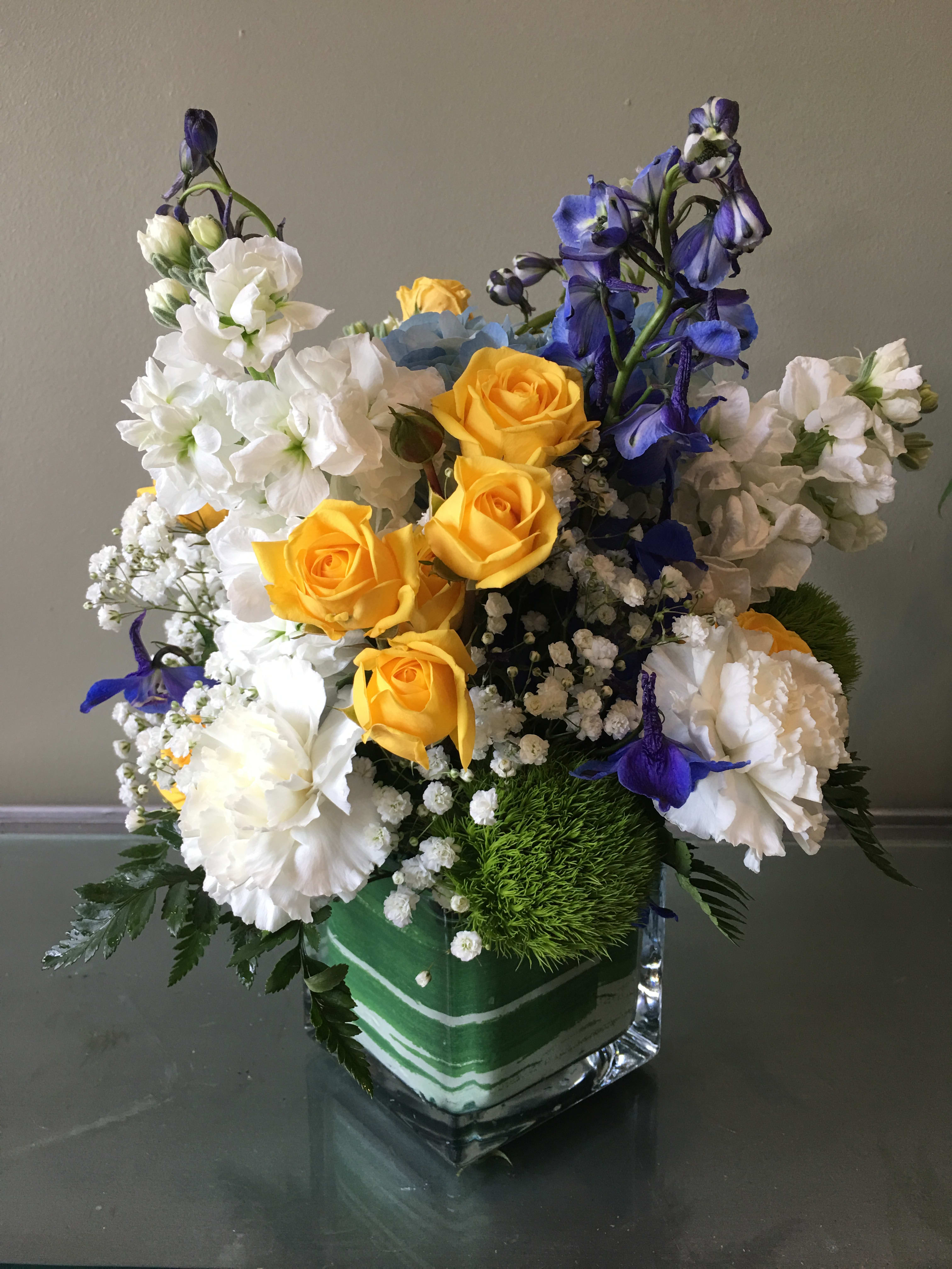 Compact arrangement of yellow roses, white blooms, and blue flowers in a square glass vase
