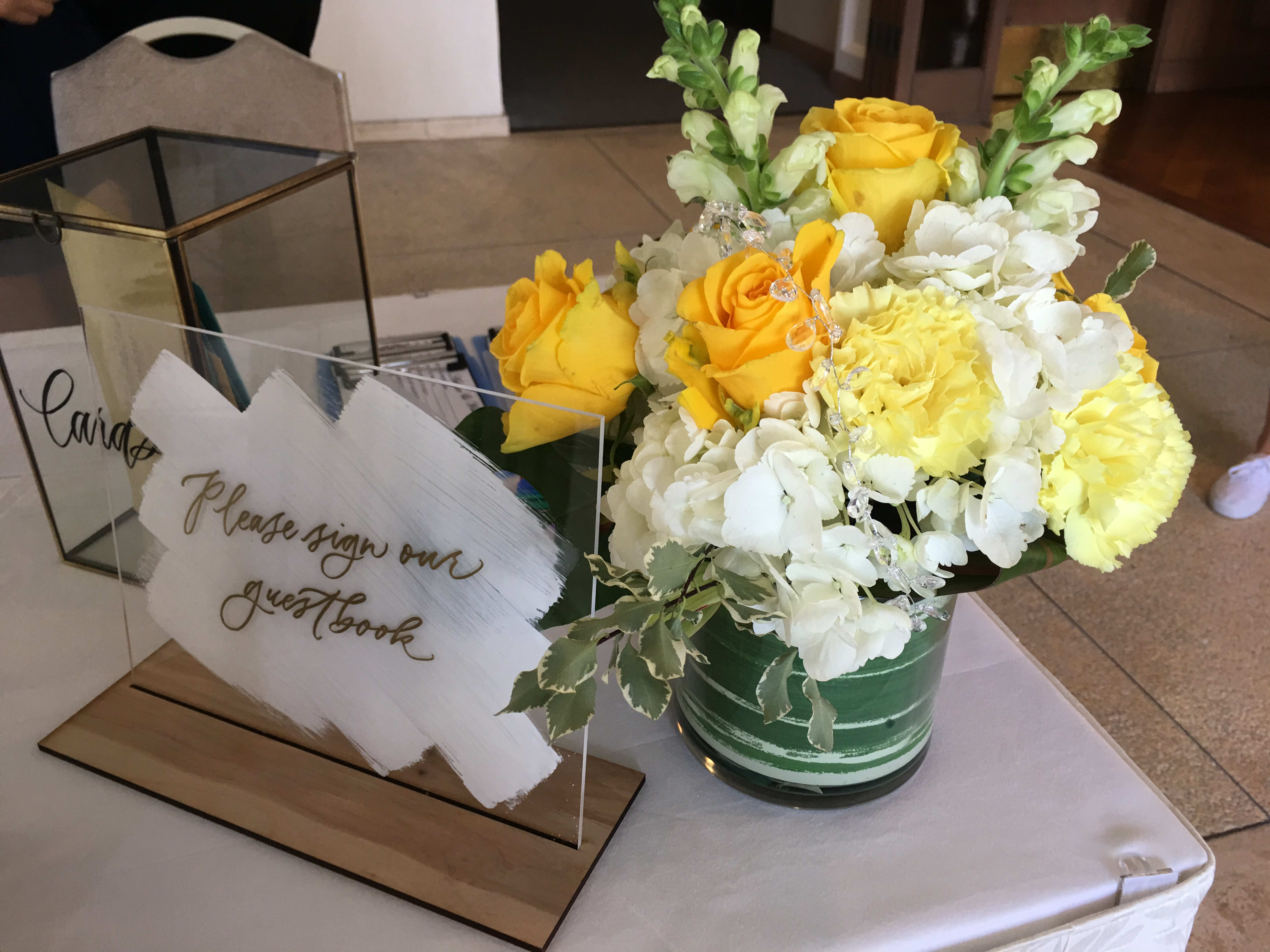 Yellow and white flower arrangement with roses and snapdragons in a green vase beside an acrylic guestbook sign