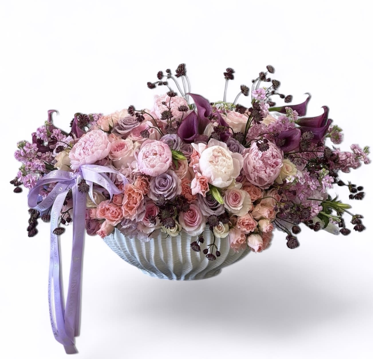 Petal Poetry - A lush, garden-style arrangement designed in a sculpted white ceramic bowl, featuring layers of premium blooms in soft blush, dusty lavender, and deep plum tones. This piece is full, airy, and intentionally unstructured—bringing a romantic, high-end feel to any space. Finished with delicate ribbon detailing for an elevated signature touch.