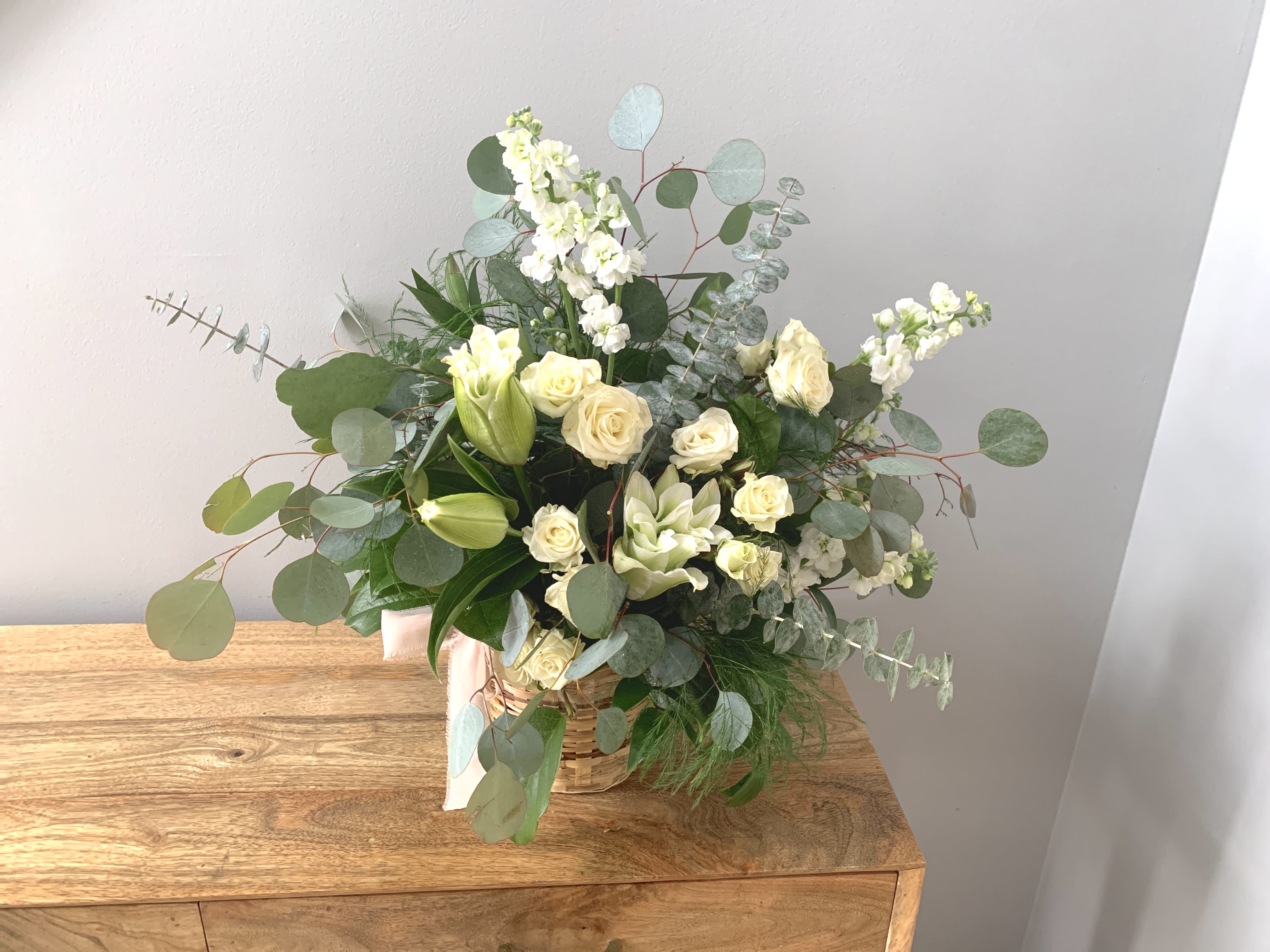 White and cream flower arrangement with roses and lilies in a woven basket with eucalyptus accents.