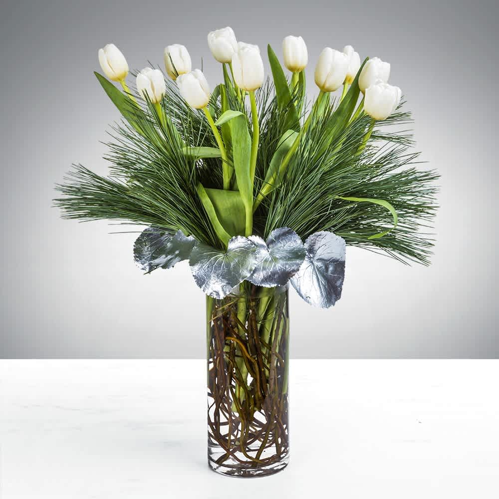 Tall arrangement of white tulips with pine and silver accents in a clear glass cylinder vase