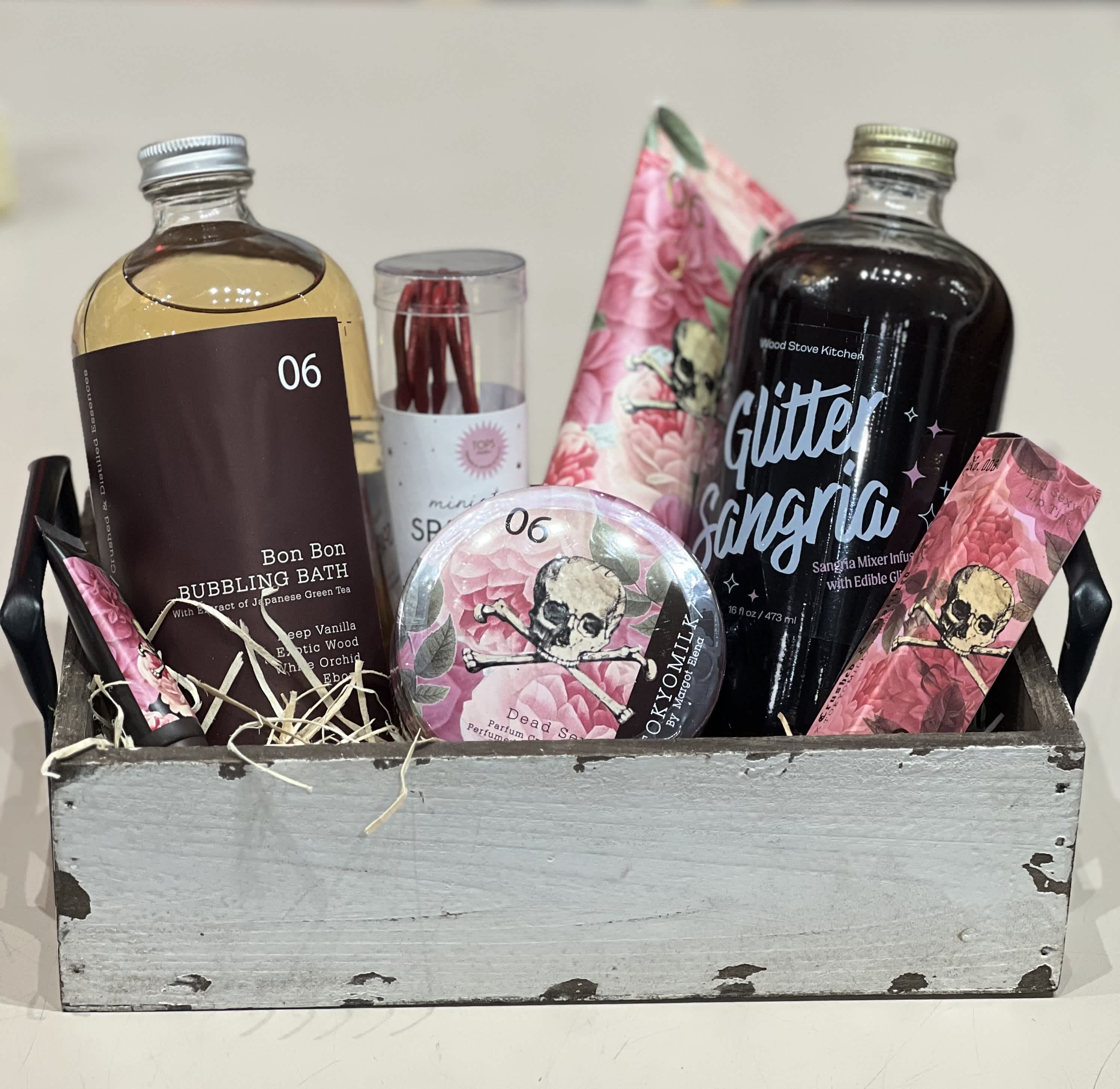 Wooden crate gift set with bath soak, sangria mixer, and pink floral skull-themed self-care items.