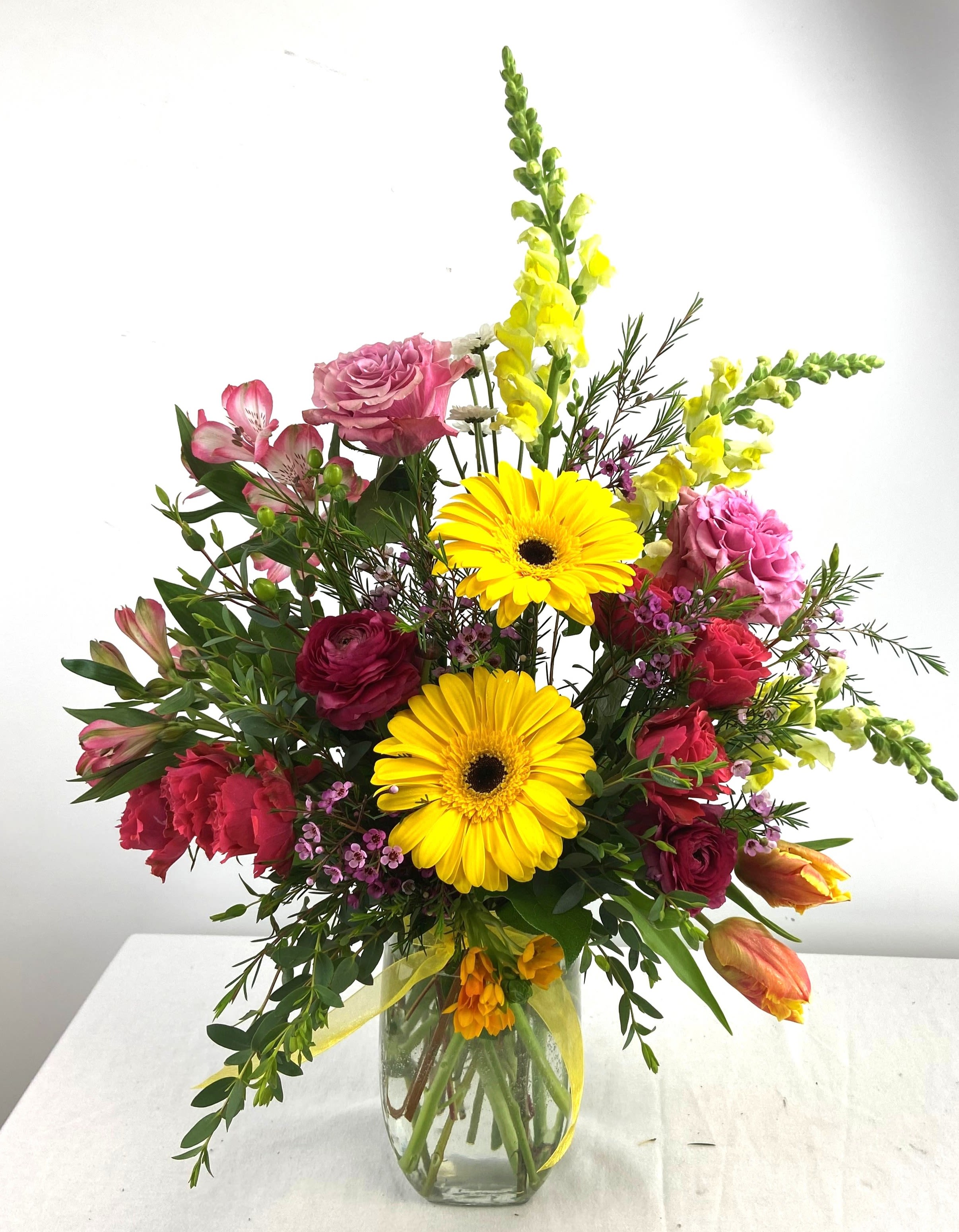 Tall mixed bouquet of yellow gerbera daisies, pink roses, and bright blooms in a clear glass vase.