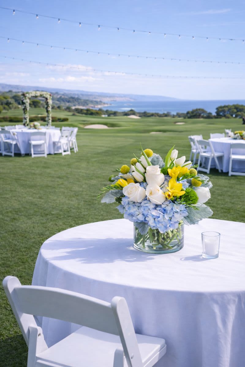 Low blue, white, and yellow flower centerpiece in a clear vase on an outdoor table with white linens