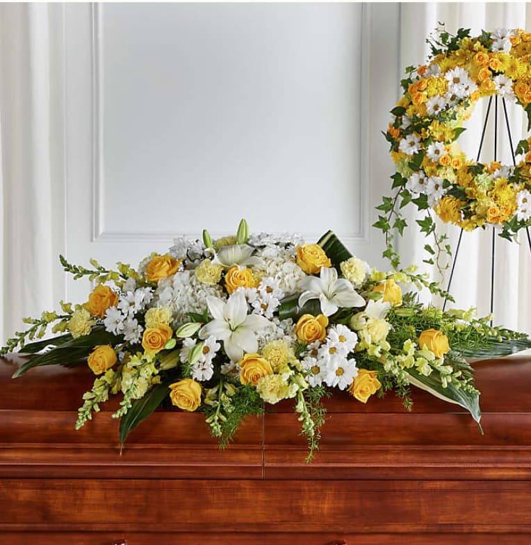 Yellow and white casket spray with roses, lilies, and daisies on a wood casket with a matching wreath behind it