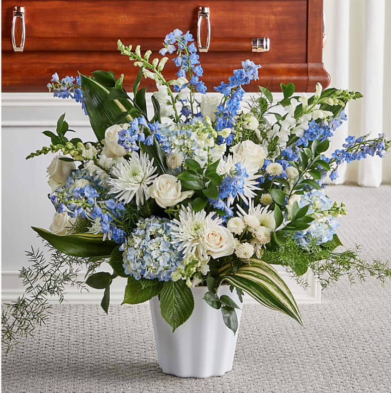 Large blue and white flower arrangement in a white urn in front of a wooden casket