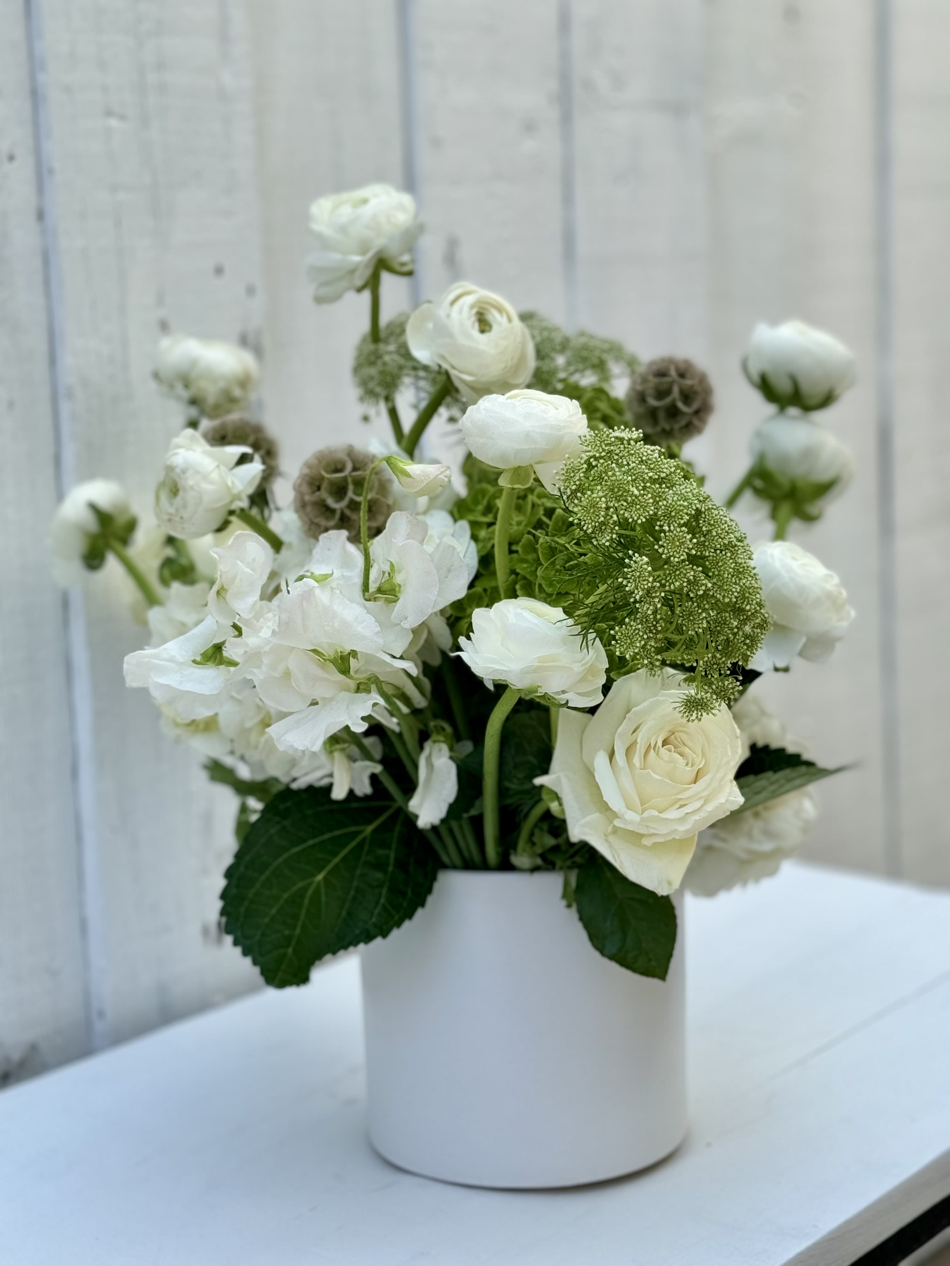White roses and ranunculus with green accent blooms arranged in a white cylinder vase.