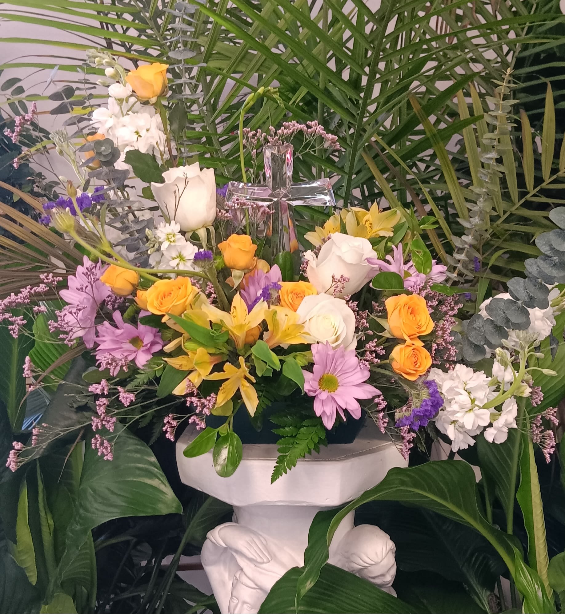 Arrangement of yellow and white flowers with a glass cross, set among tall palm greenery on a white pedestal.