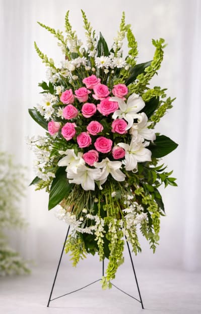 Graceful Pink Tribute Spray - A sculptural standing spray that speaks with quiet dignity-this flower arrangement is crafted as a tall memorial spray on a black metal easel, ideal for a funeral service or graveside tribute. The design centers on a dense cluster of bright pink roses and is framed by large white lilies and smaller white daisy-like blooms, creating a compassionate, heartfelt statement for a loved one or to mark a solemn occasion.  Soft cascades of Bells of Ireland and delicate white accent flowers give the piece graceful downward movement and layered texture, while the mix of pink, white, and green provides a balanced, serene palette. Measures as a full vertical spray on a sturdy easel for prominent display; the combination of long-stemmed lilies, snapdragons, roses, and daisies ensures visual impact from a distance and thoughtful detail up close.  Made to comfort and honor, this arrangement offers a meaningful way to say goodbye, celebrate a life, or express sympathy-she'll feel genuinely remembered when this is chosen in her honor. The spray arrives ready to stand on the included black metal easel; for lasting freshness, keep the stems supplied with water where possible and follow any care guidance provided at delivery.