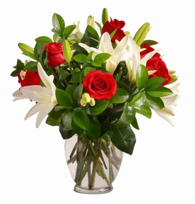Crimson &amp; Ivory Elegance - Bold red roses meet open white lilies in a romantic table arrangement perfect for anniversaries, Valentine's Day, or any moment you want to make someone feel truly cherished. The contrast of deep crimson blooms against pure white lilies creates an immediate, heart-stirring impression that reads as both celebratory and intimate - the kind of bouquet that tells the person you're thinking of them in a meaningful way.  Carefully gathered stems sit in a rounded clear glass vase, giving the design a classic upright profile that fits beautifully on a dining table, desk, or bedside. Glossy dark green foliage forms a generous collar around the roses and lilies, adding lush fullness and framing each bloom so their color and texture pop. Ideal for gifting to a partner or a loved one on a special day, this arrangement arrives ready to display; change the water regularly and trim stems every few days to help the roses and lilies stay fresh longer.