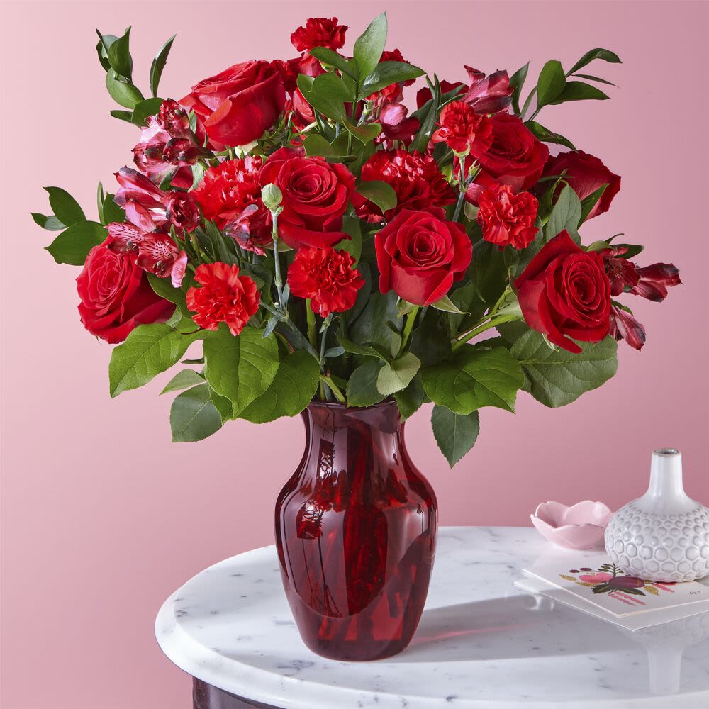 Arrangement of red roses and carnations in a red glass vase on a marble table.