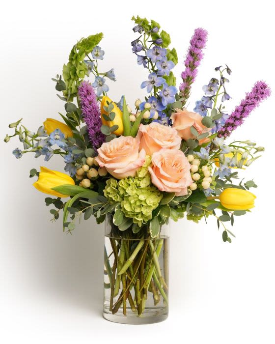 Mixed bouquet of peach roses, yellow tulips, purple and blue blooms in a clear glass vase