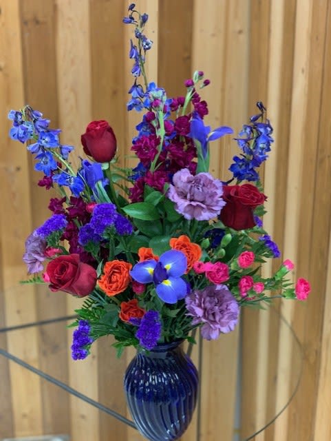 Tall mixed arrangement with red roses, purple delphinium and carnations in a blue glass vase