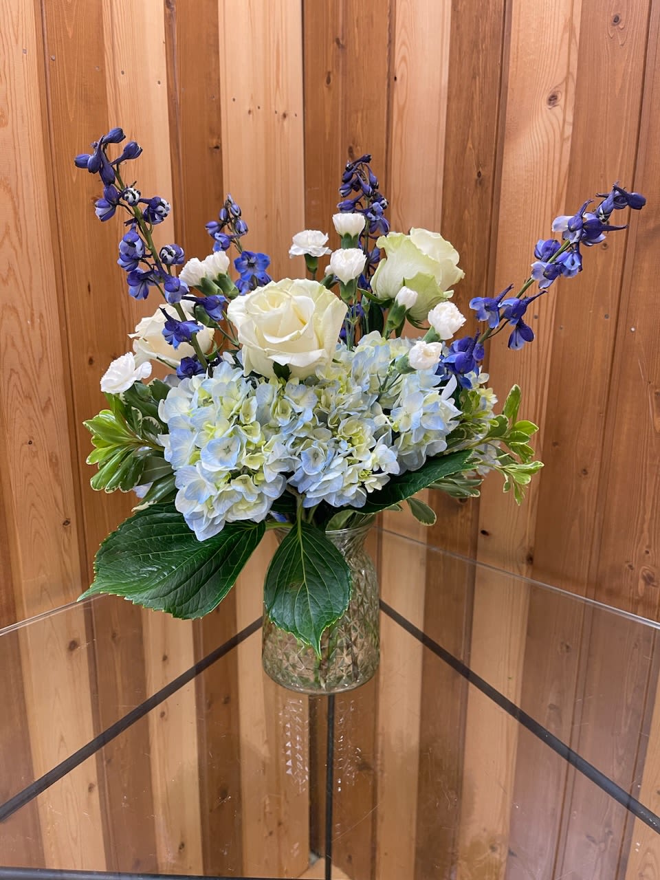 Blue hydrangeas with white roses and tall blue delphinium in a clear patterned glass vase