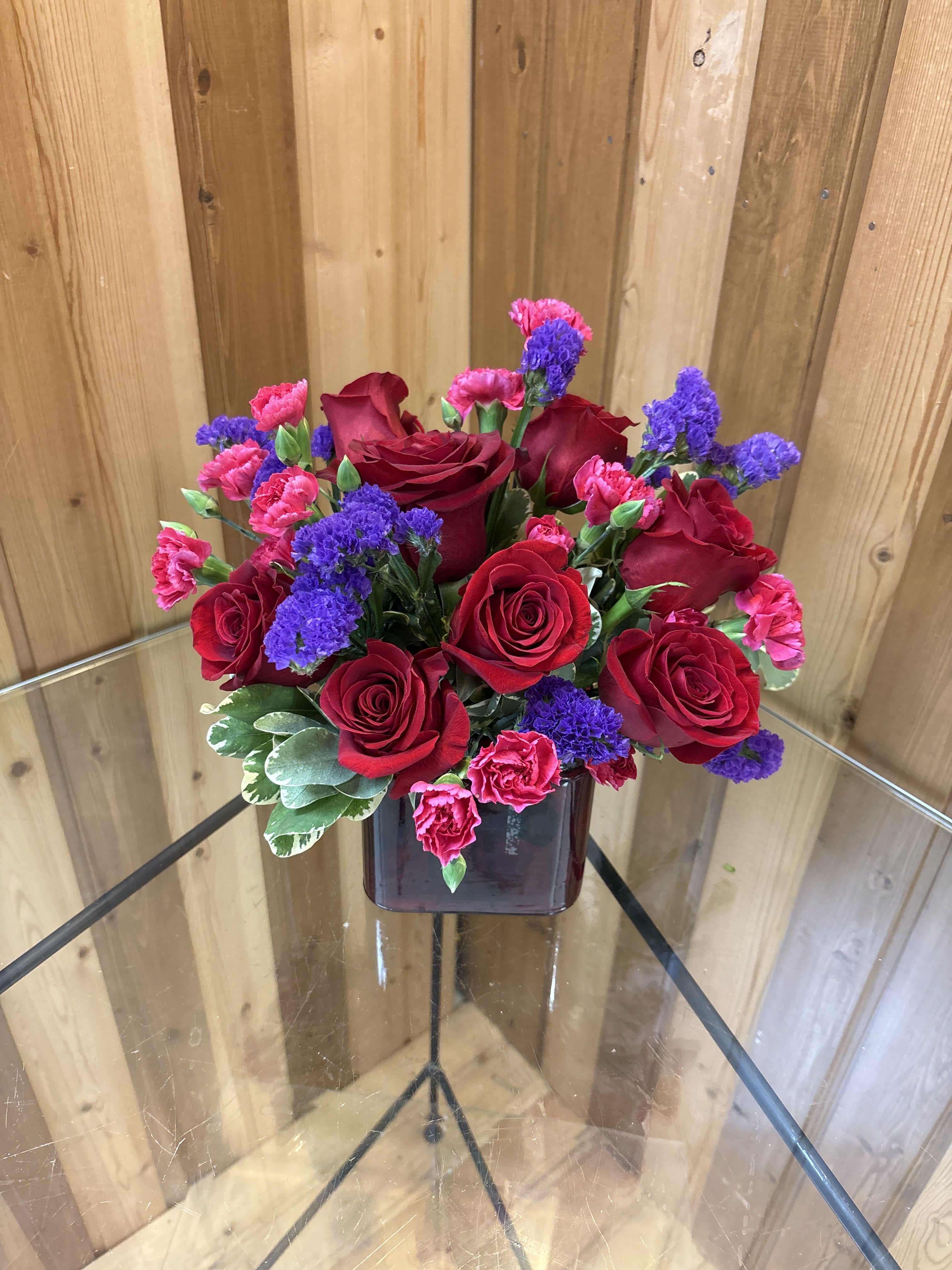 Red roses, pink carnations, and purple flowers arranged in a dark red cube vase