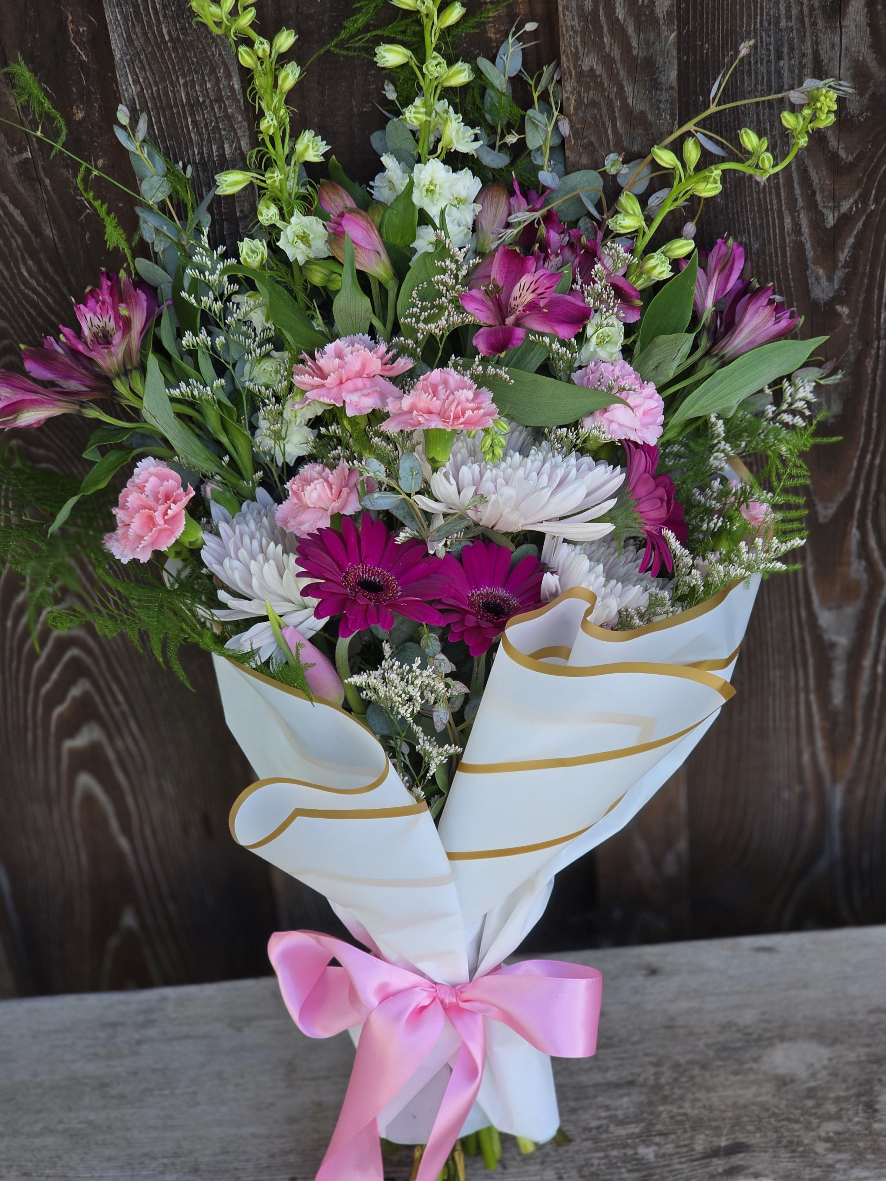 Hand-tied bouquet of pink and white flowers wrapped in white paper with a pink bow.